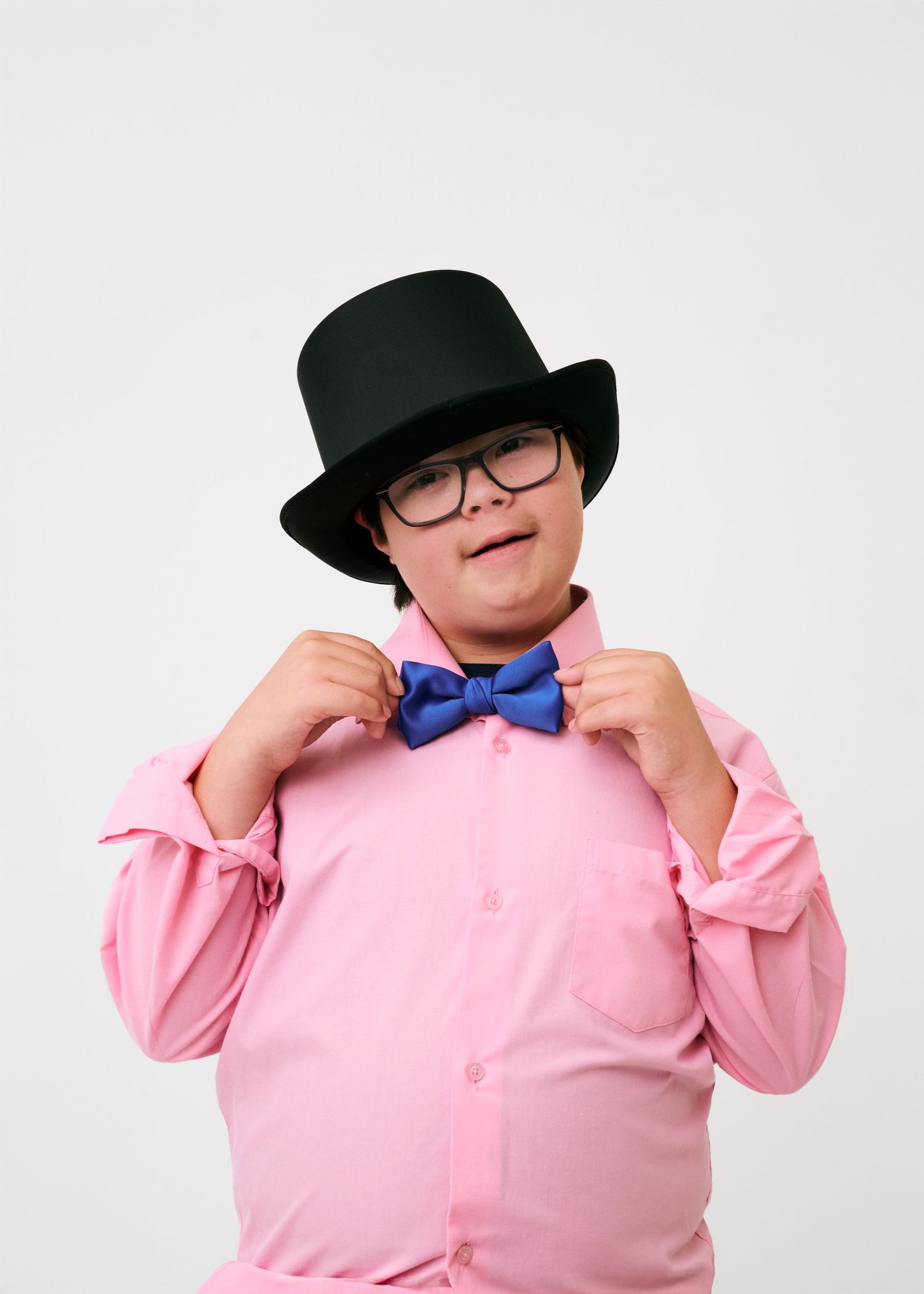 Person wearing pink button-up shirt, black hat and blue bowtie poses playfully against white background.