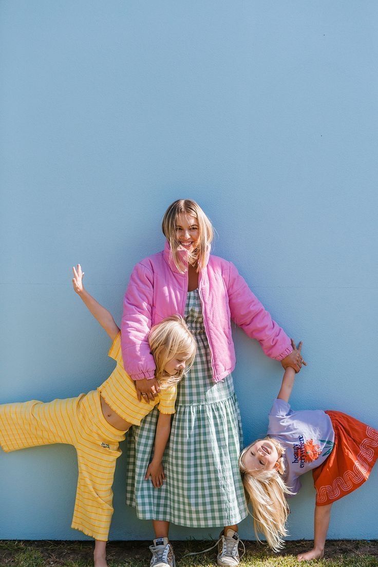 Woman in pink jacket poses with two playful blonde children against a bright blue wall outdoors.