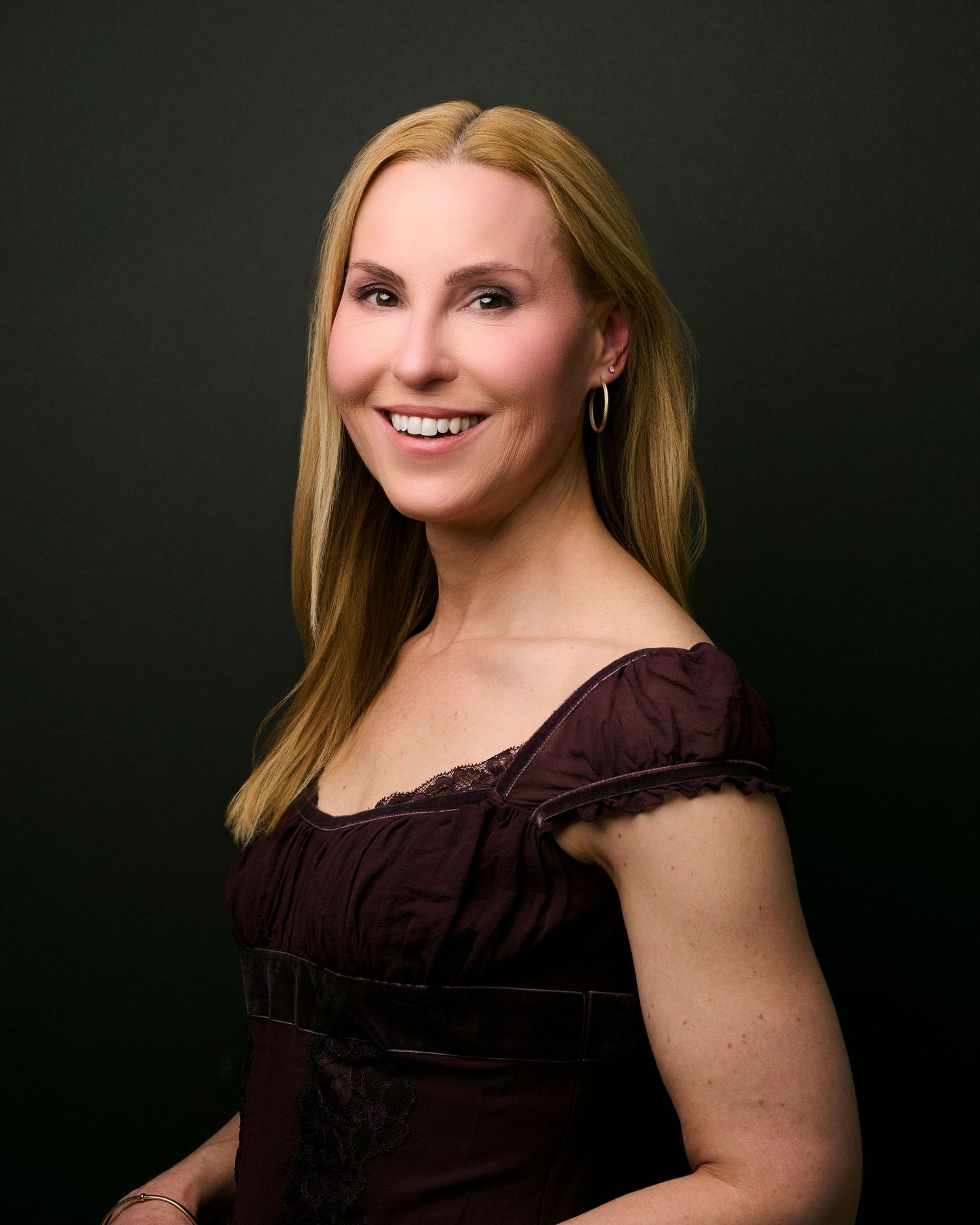 Professional studio portrait against dark background showing a woman in burgundy ruffled top with blonde hair and warm smile.