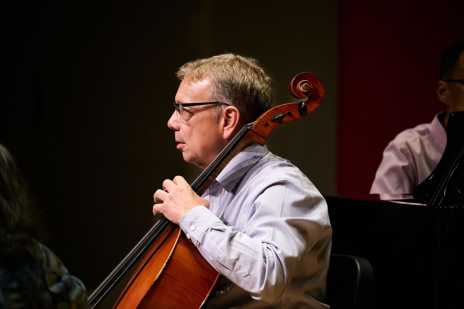 Man with glasses playing cello during a musical performance on a dimly lit stage.