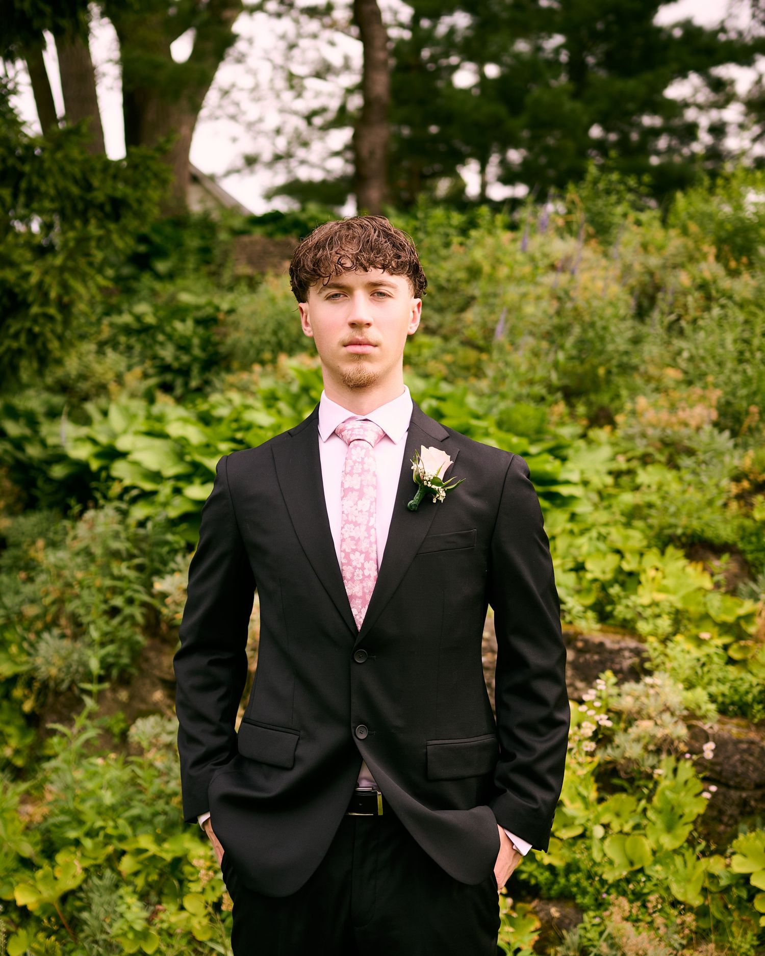 Young man in black suit with pink tie and boutonniere poses in lush garden setting.