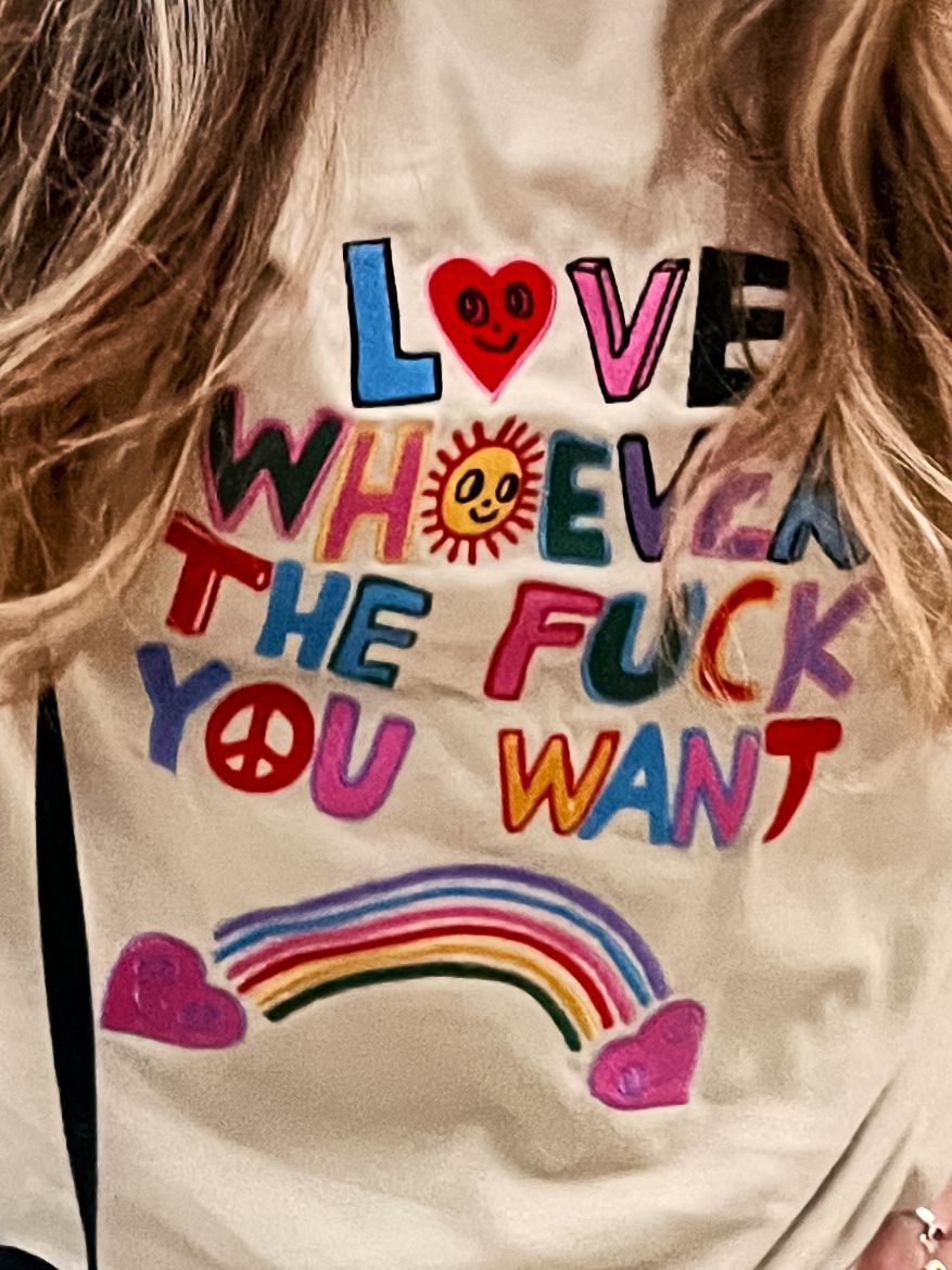 Colorful lettering spells out 'Love whoever the fuck you want' with a rainbow and heart on a light colored t-shirt.