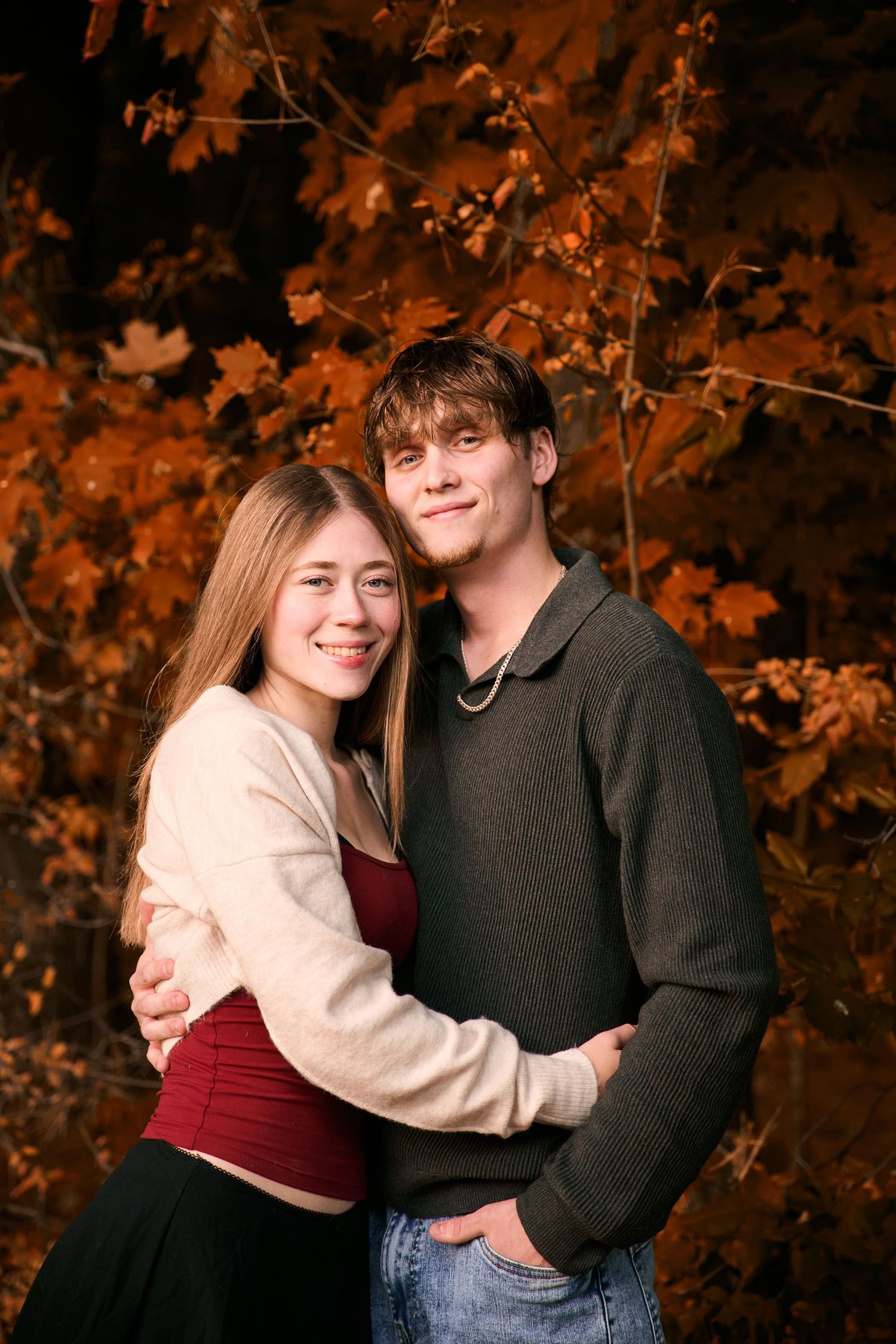 A couple embraces while posing for an autumn portrait with orange fall foliage in the background.