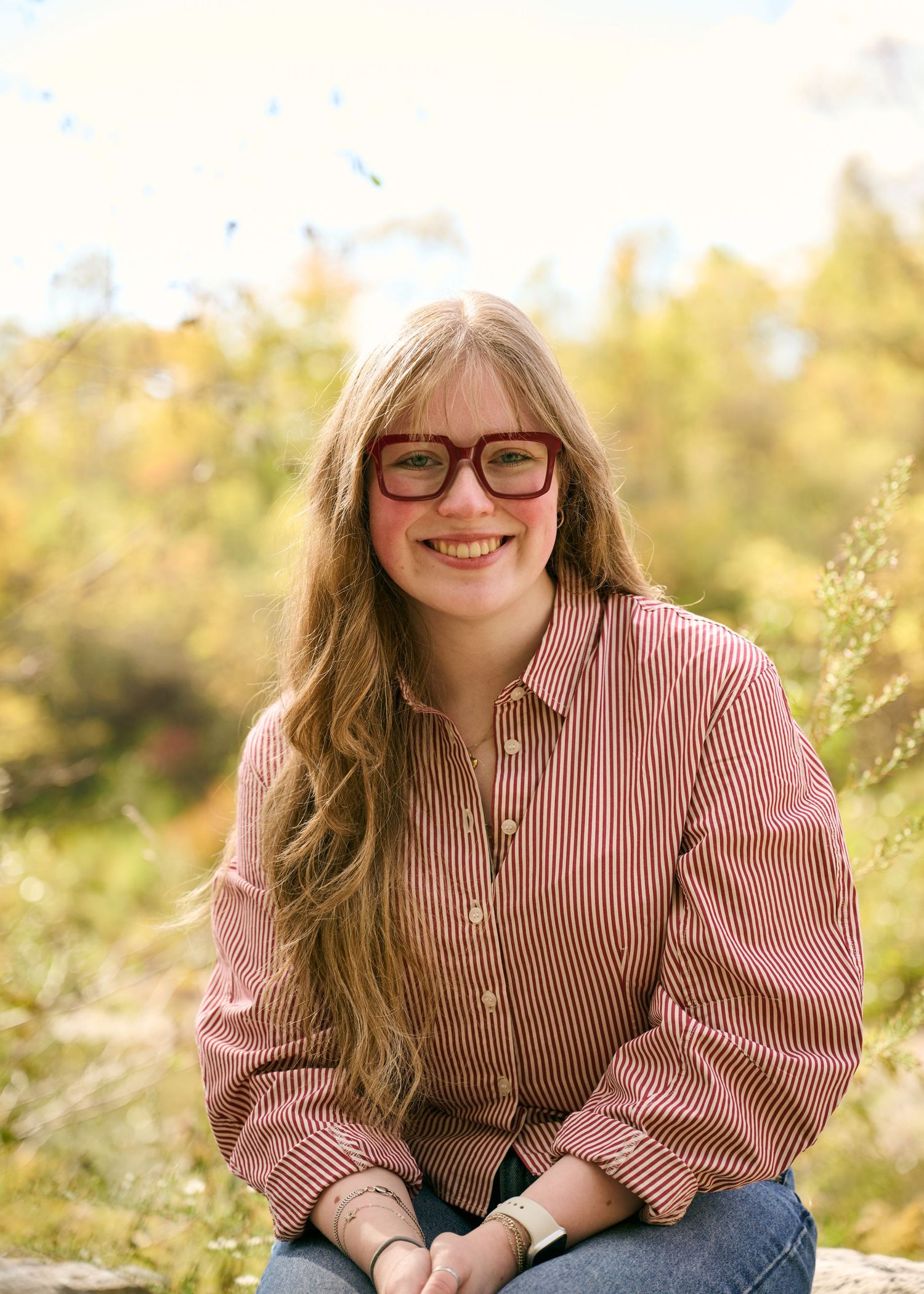 A person wearing glasses and a red striped button-down shirt smiles outdoors on a sunny autumn day.