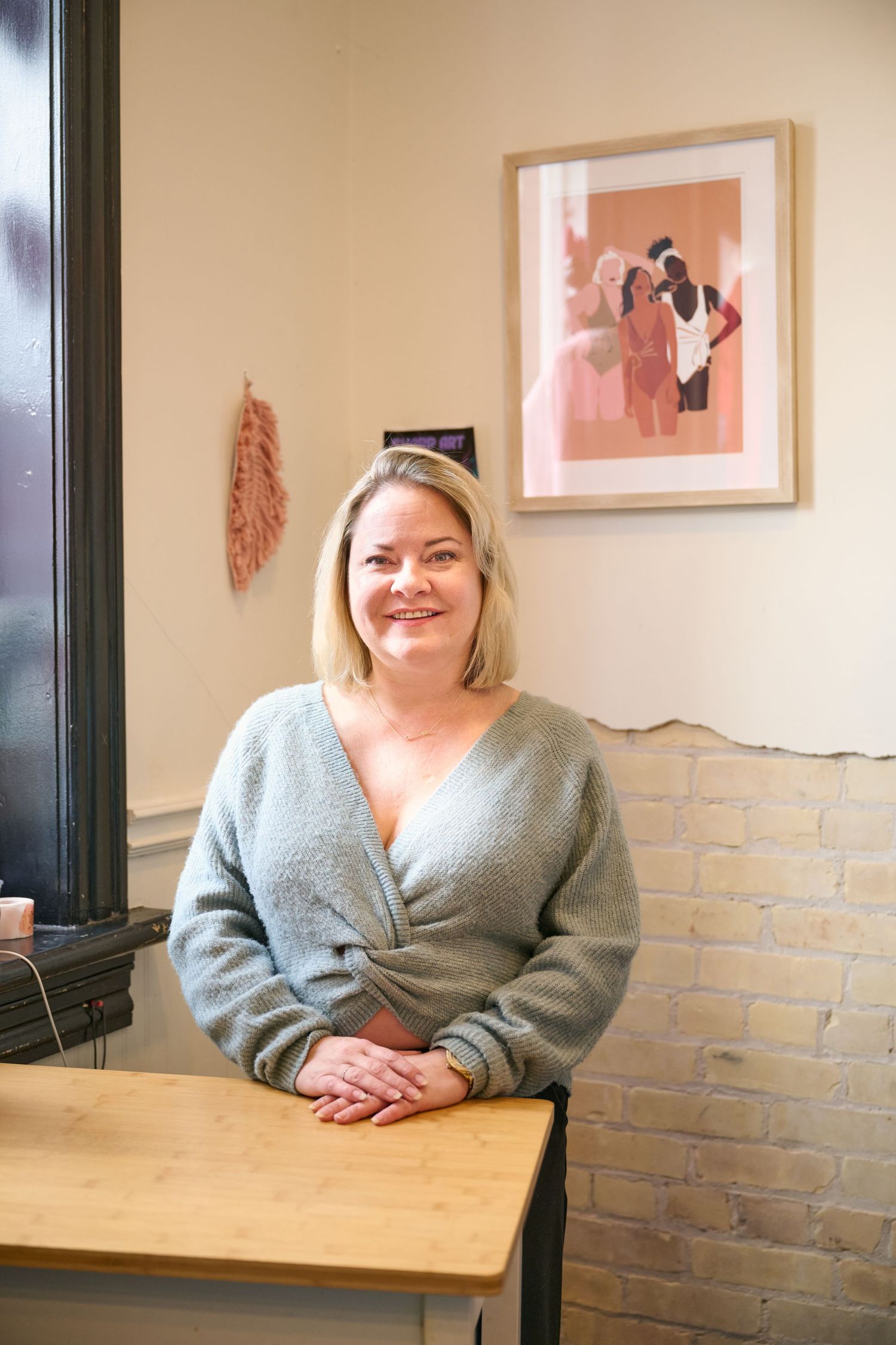 Person in gray sweater sitting at wooden table with framed artwork on beige wall behind them.