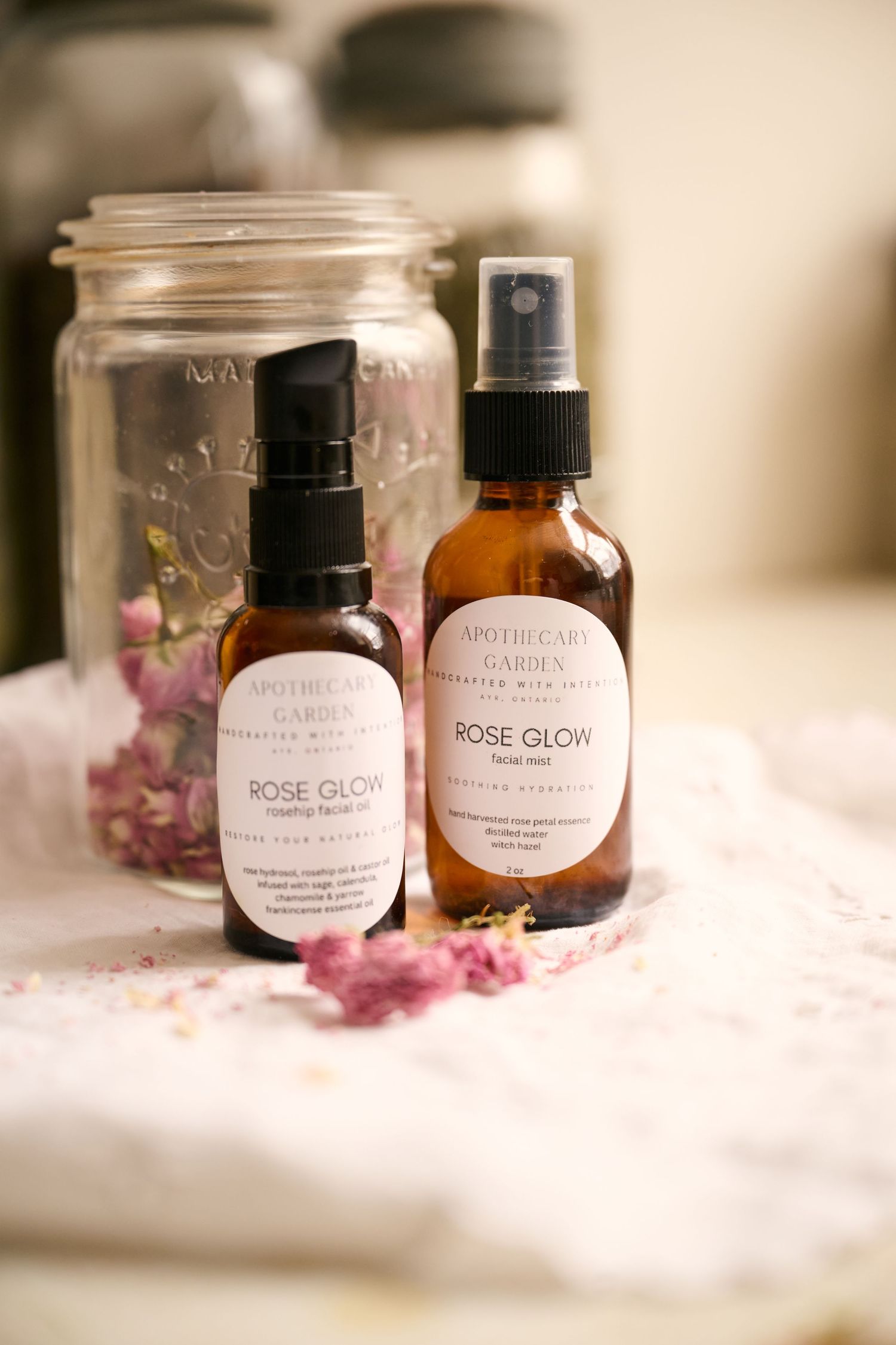 Two bottles of Rose Glow beauty product with dried pink flowers and a mason jar on a soft white surface.