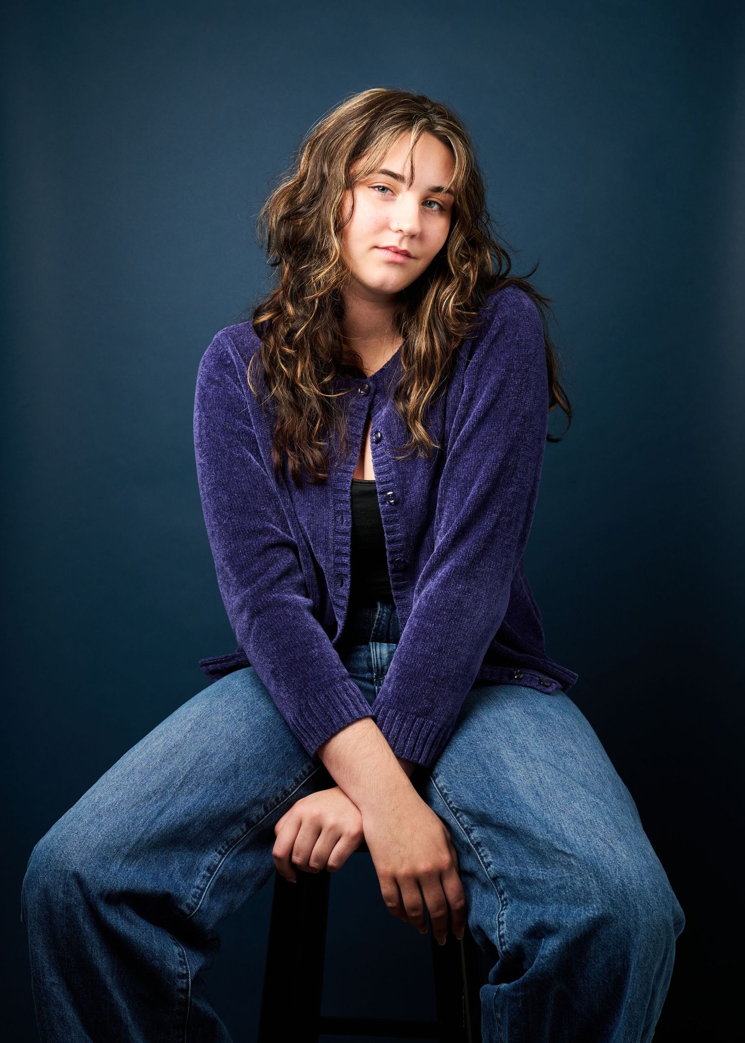 Person in blue jeans and purple sweater sits casually on dark background in fashion photoshoot series.
