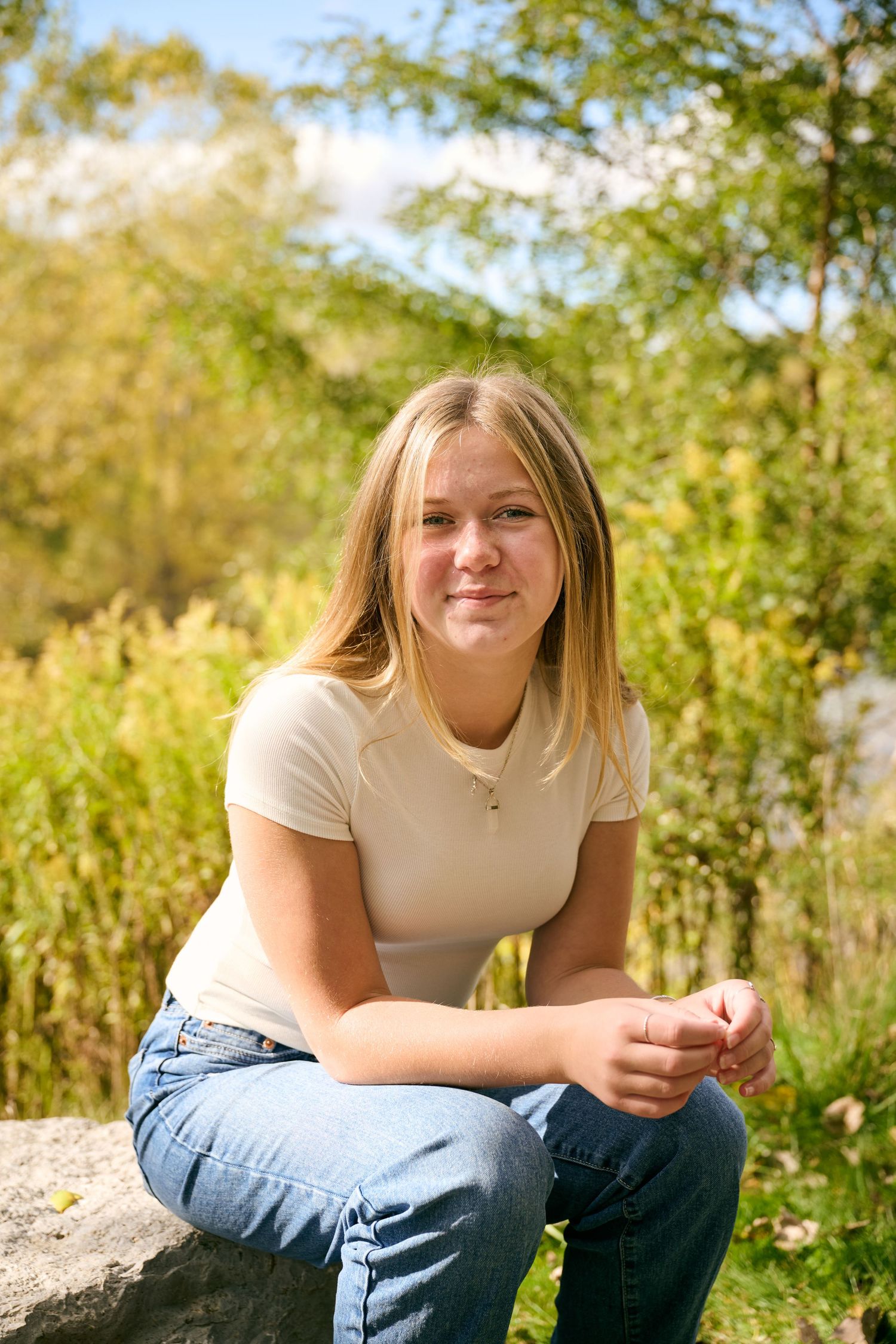 Person in light colored shirt and jeans sits casually outdoors on sunny day with greenery in background.