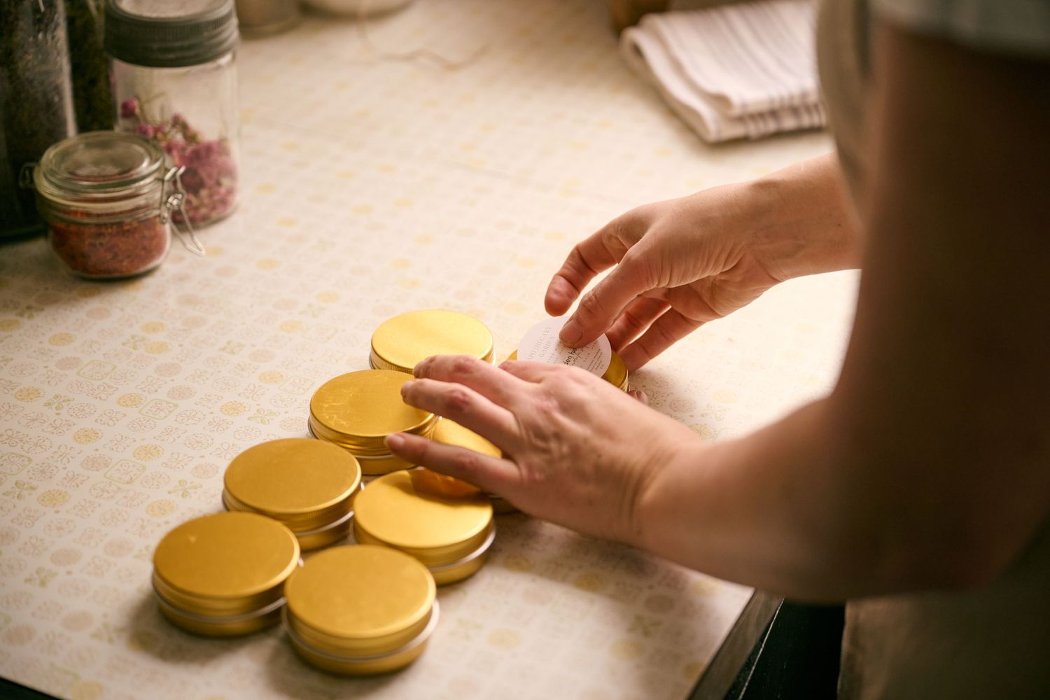 Hands arranging rows of golden metal jar lids on a white countertop in warm lighting.