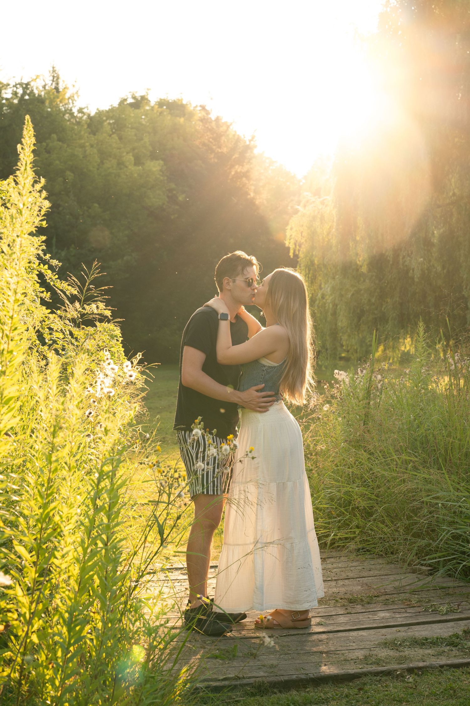 Romantic couple standing together on wooden dock at sunset with glowing golden light and greenery in background.