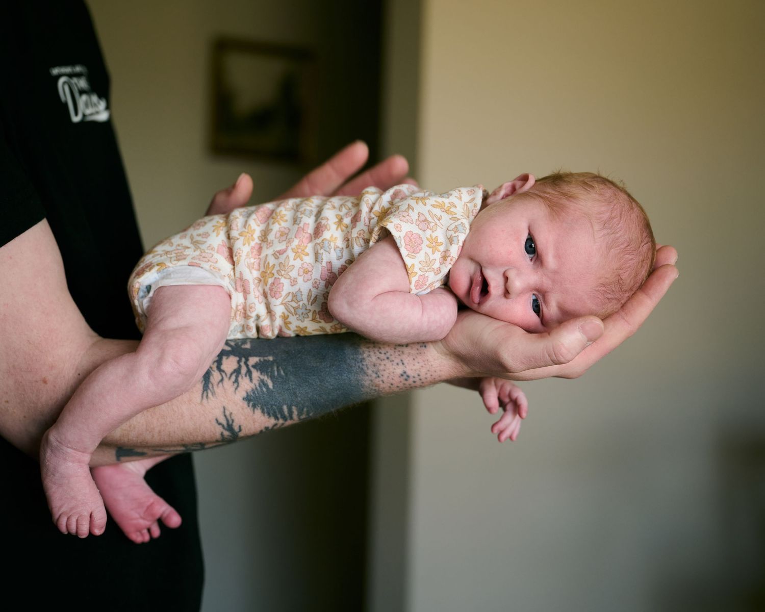 Tattooed adult hands cradling a tiny newborn baby in a floral onesie, gently supporting the infant horizontally.