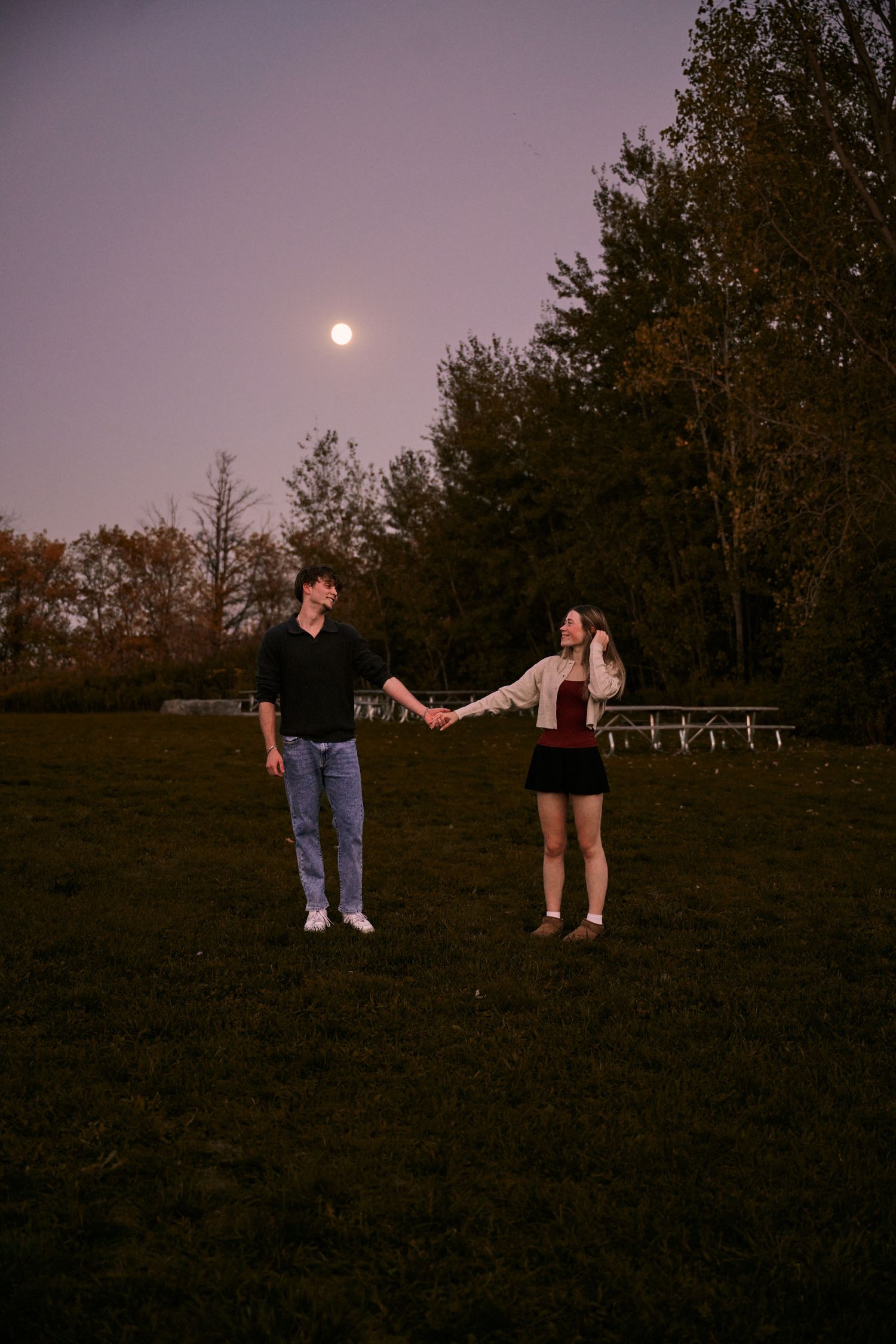 A couple holds hands in a grassy field at dusk with a full moon glowing in the purple sky.