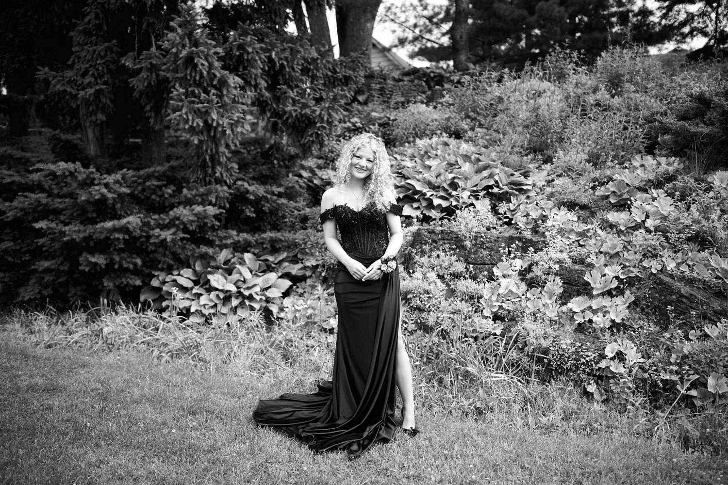 Woman in elegant black off-shoulder gown posing in lush garden, black and white photo.