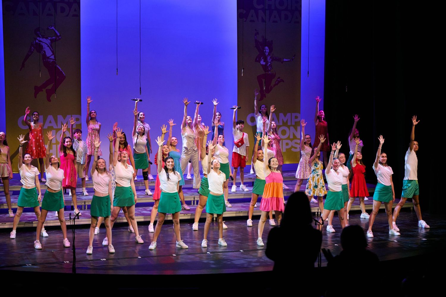 Dance performers in colorful costumes perform a synchronized routine on stage under bright blue lighting.
