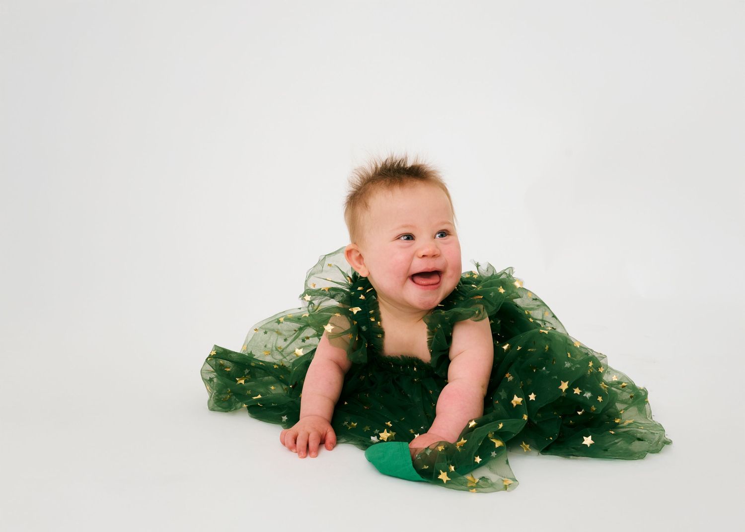 Happy baby wearing green tulle dress lies on white backdrop while smiling during professional photoshoot session.