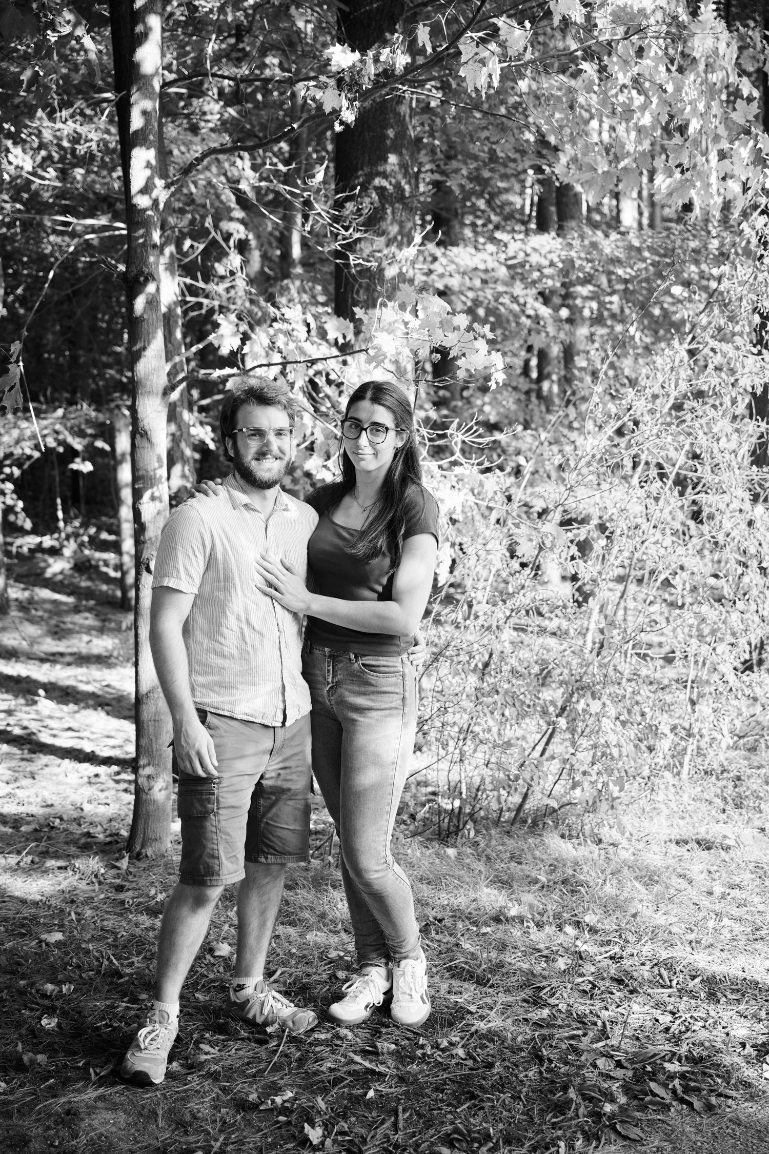 A couple stands together on a wooded trail in black and white photography.