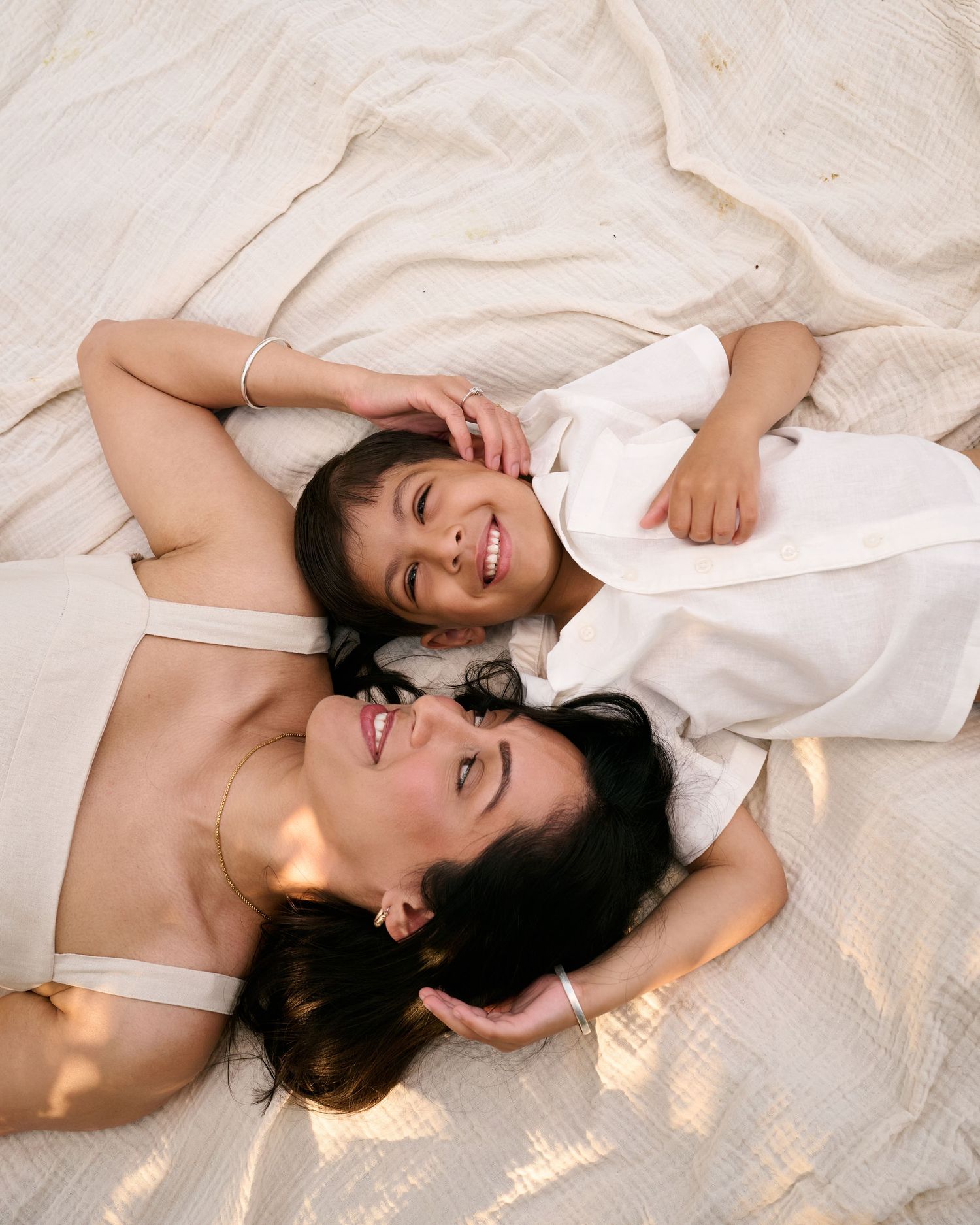 A woman and young child lying on a white bed, smiling and enjoying a tender moment together.