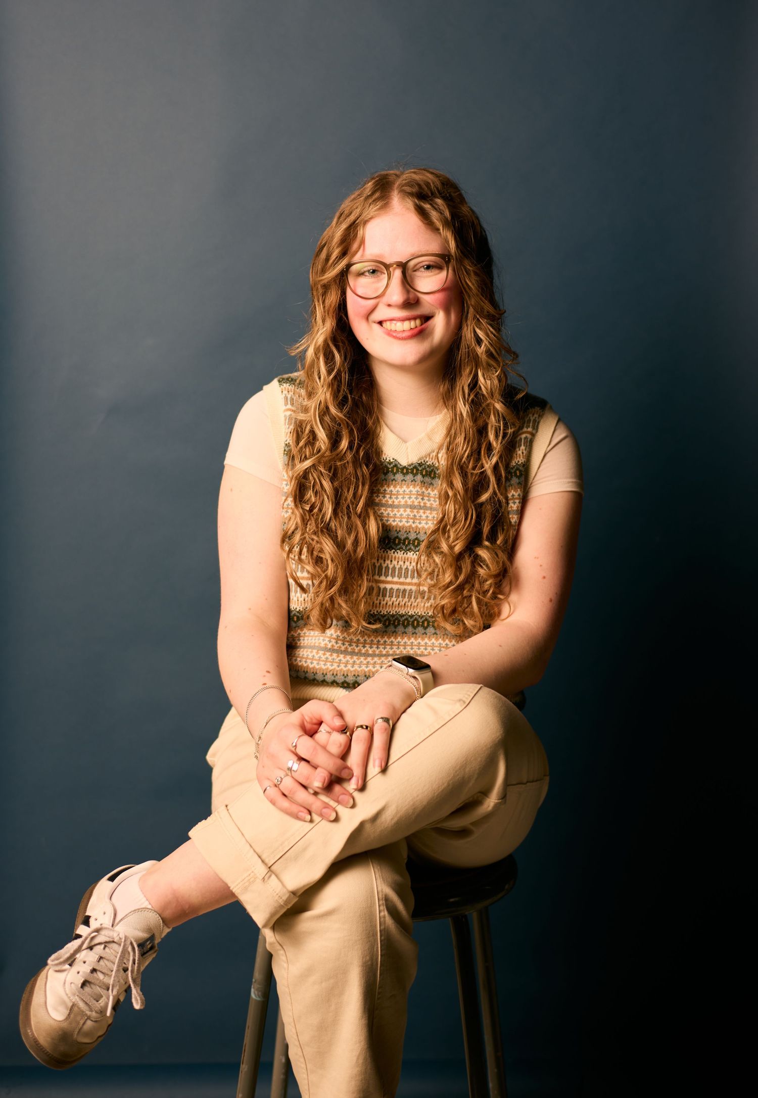 A studio portrait featuring curly honey-blonde hair, glasses, and a striped sweater against a dark backdrop.