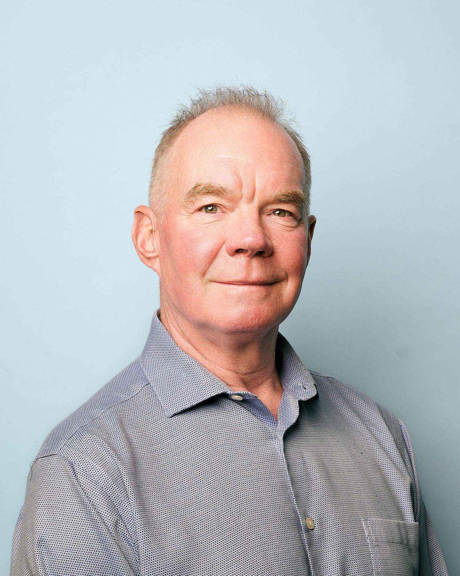 Professional headshot of a senior man with short gray hair wearing a light blue collared shirt against a blue background.