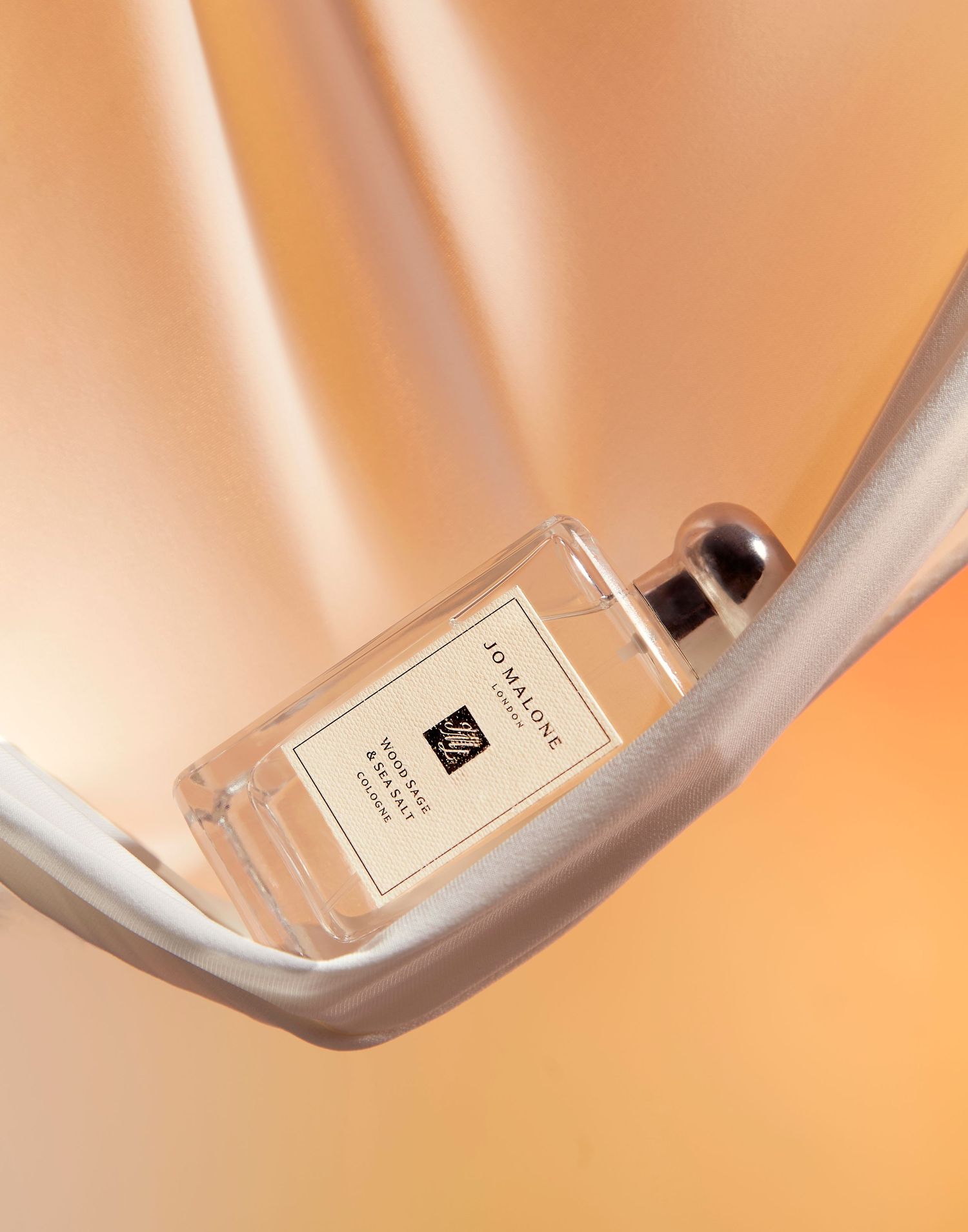 Luxury perfume bottle balanced on curved silver surface against peachy orange background.