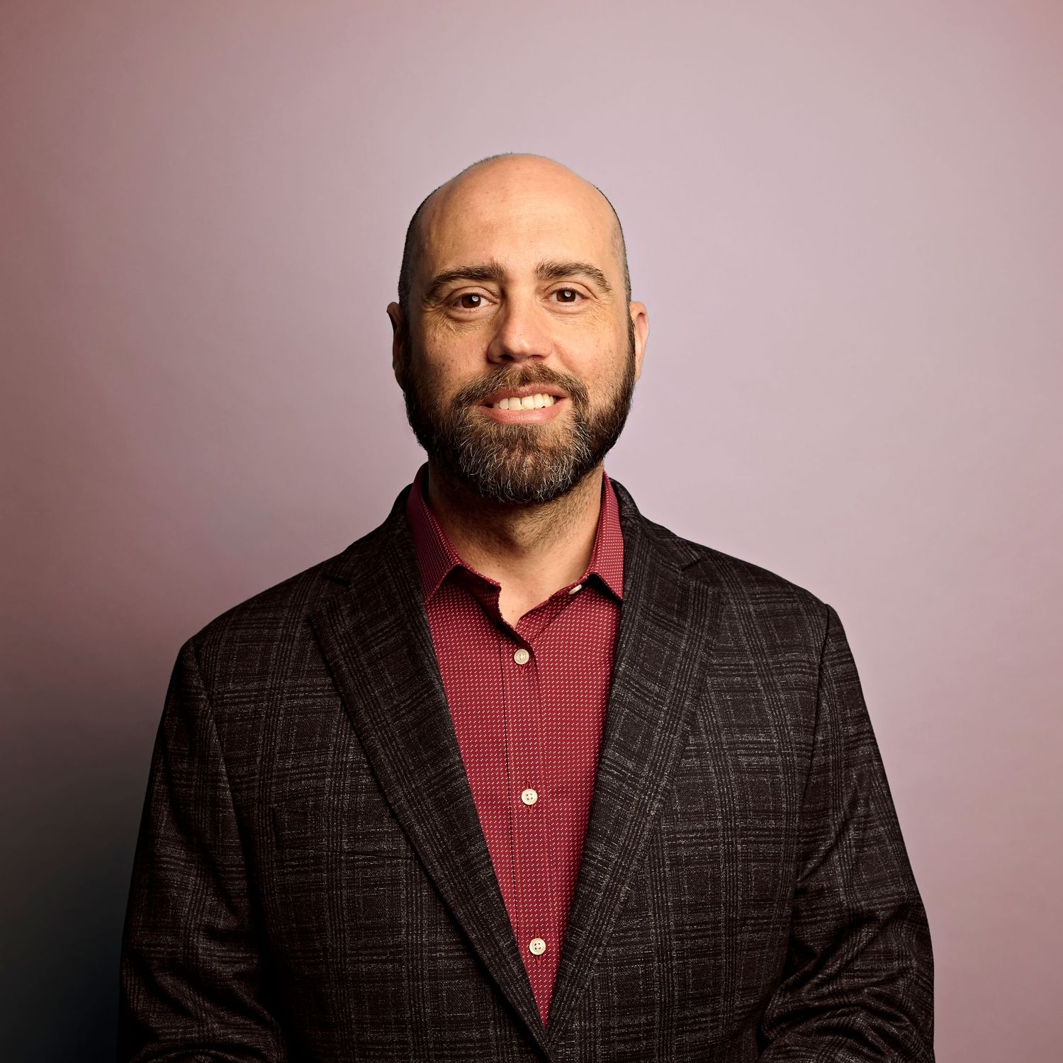 Professional headshot of a smiling man with a beard wearing a burgundy shirt and dark plaid blazer against pink background.