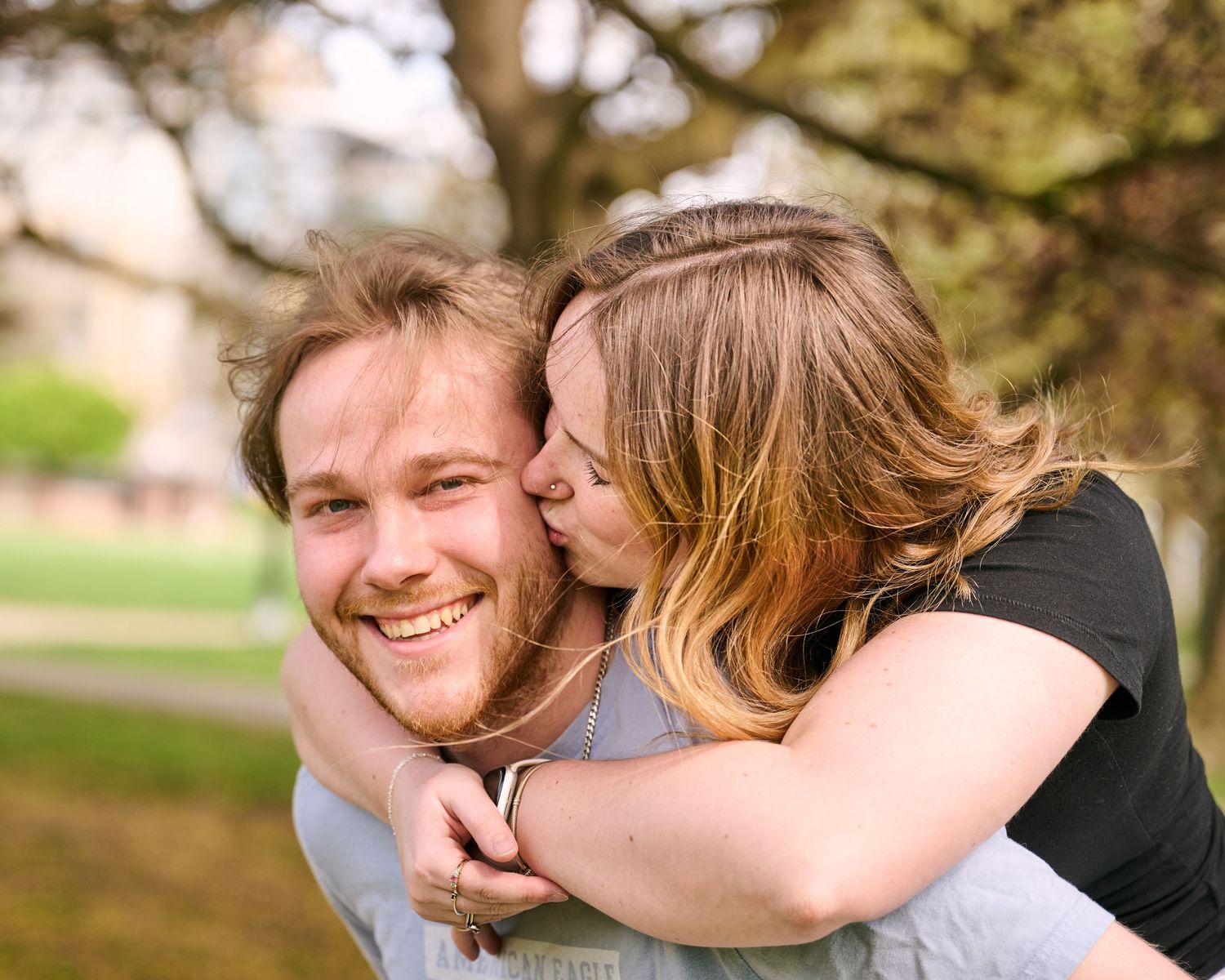 Happy couple outdoors, woman kissing smiling bearded man on cheek during piggyback ride in green park.