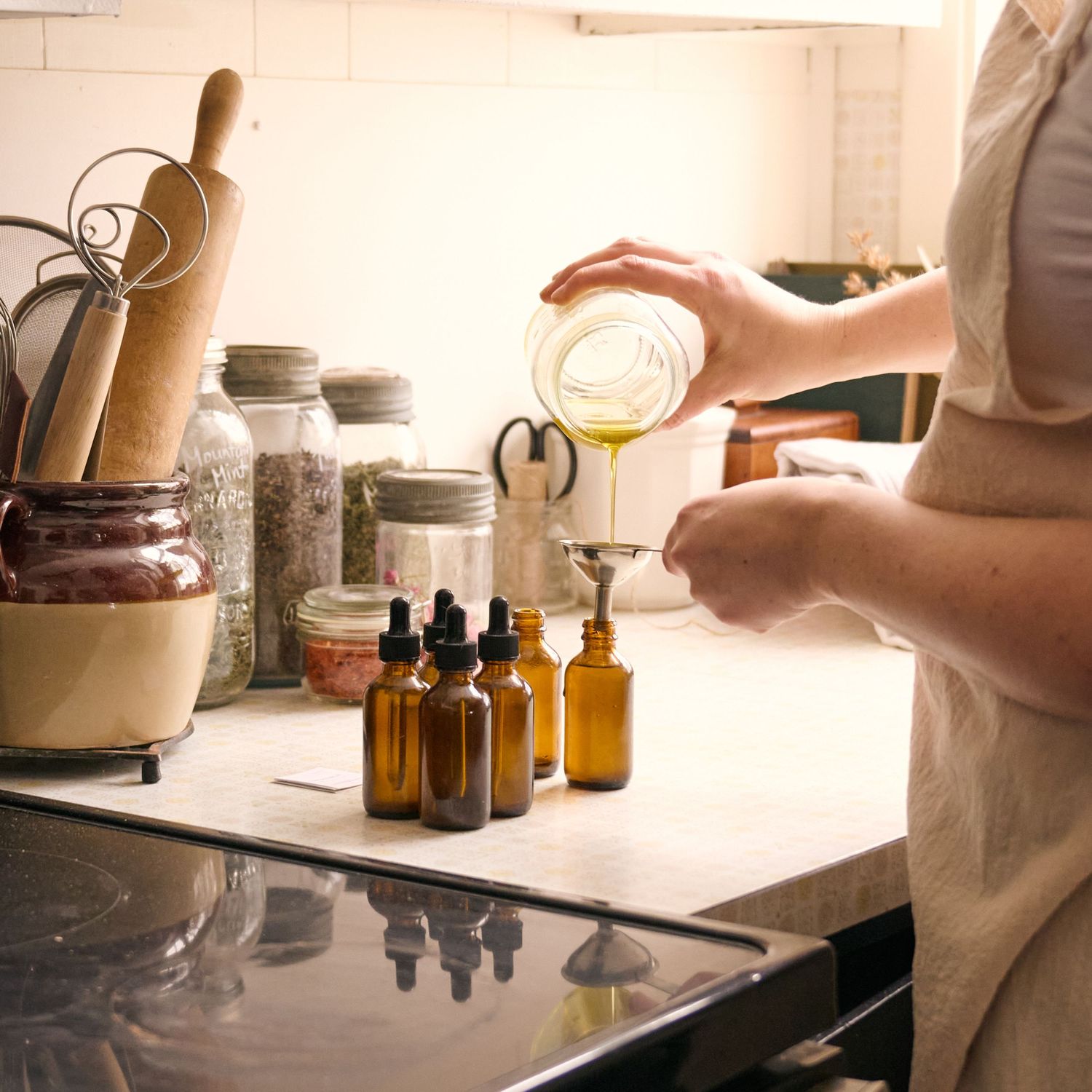 A person wearing white apron pours liquid into amber glass bottles on kitchen counter with ingredients and equipment.