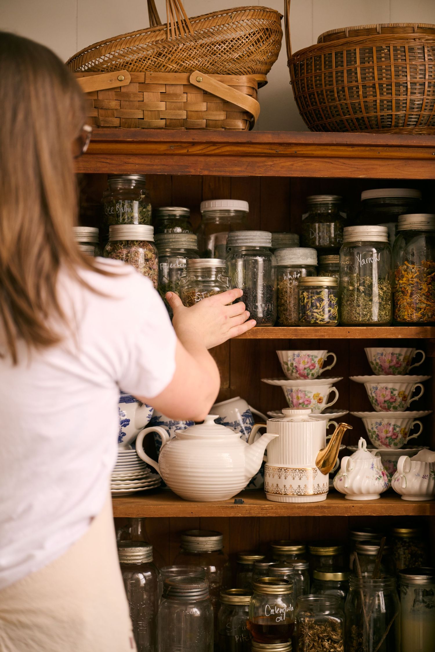 Wooden shelves displaying an organized collection of glass jars, teapots, teacups and woven baskets in a cozy pantry.