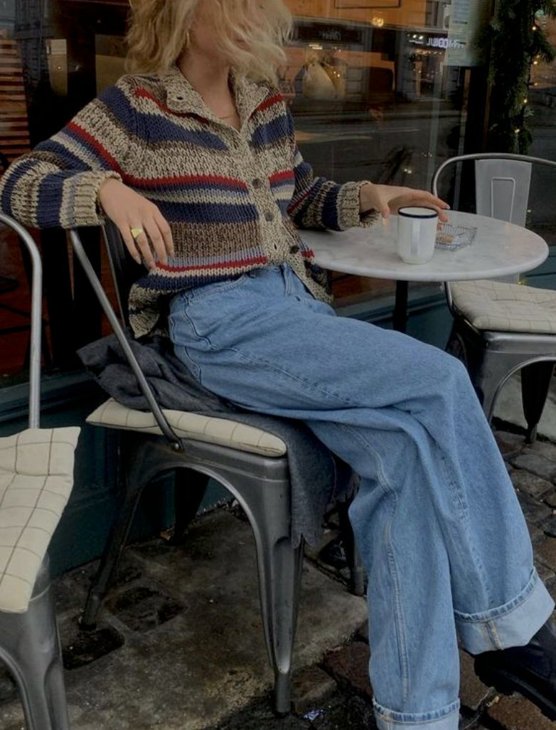 Person in striped sweater and jeans sits casually at outdoor cafe table with coffee cup.