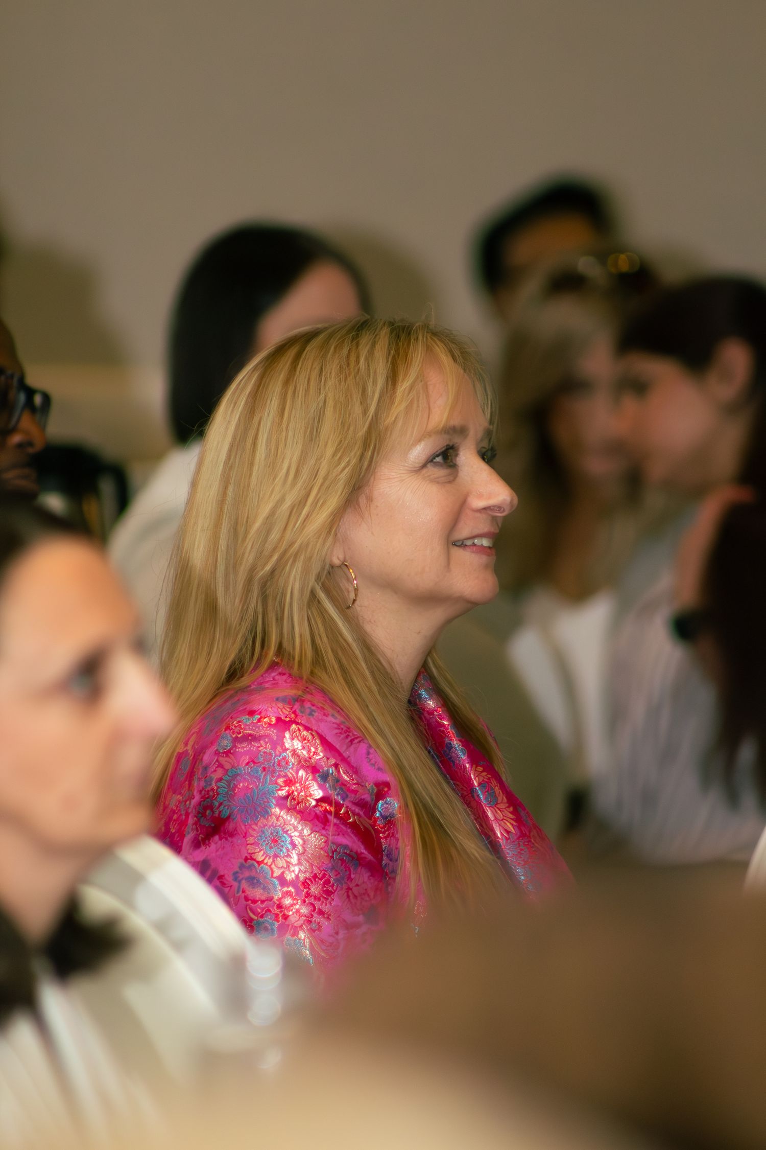 Woman in bright pink floral outfit smiles while seated in audience at indoor event.