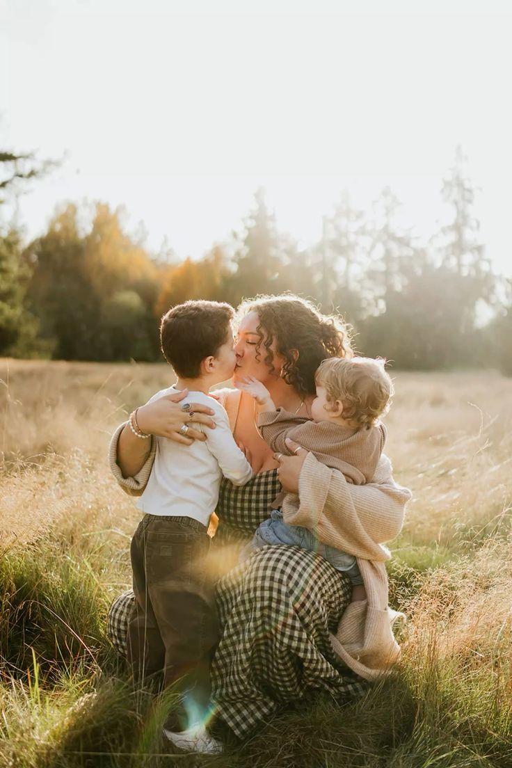 A mother kisses her young son while holding a toddler in a sunlit golden field at sunset.