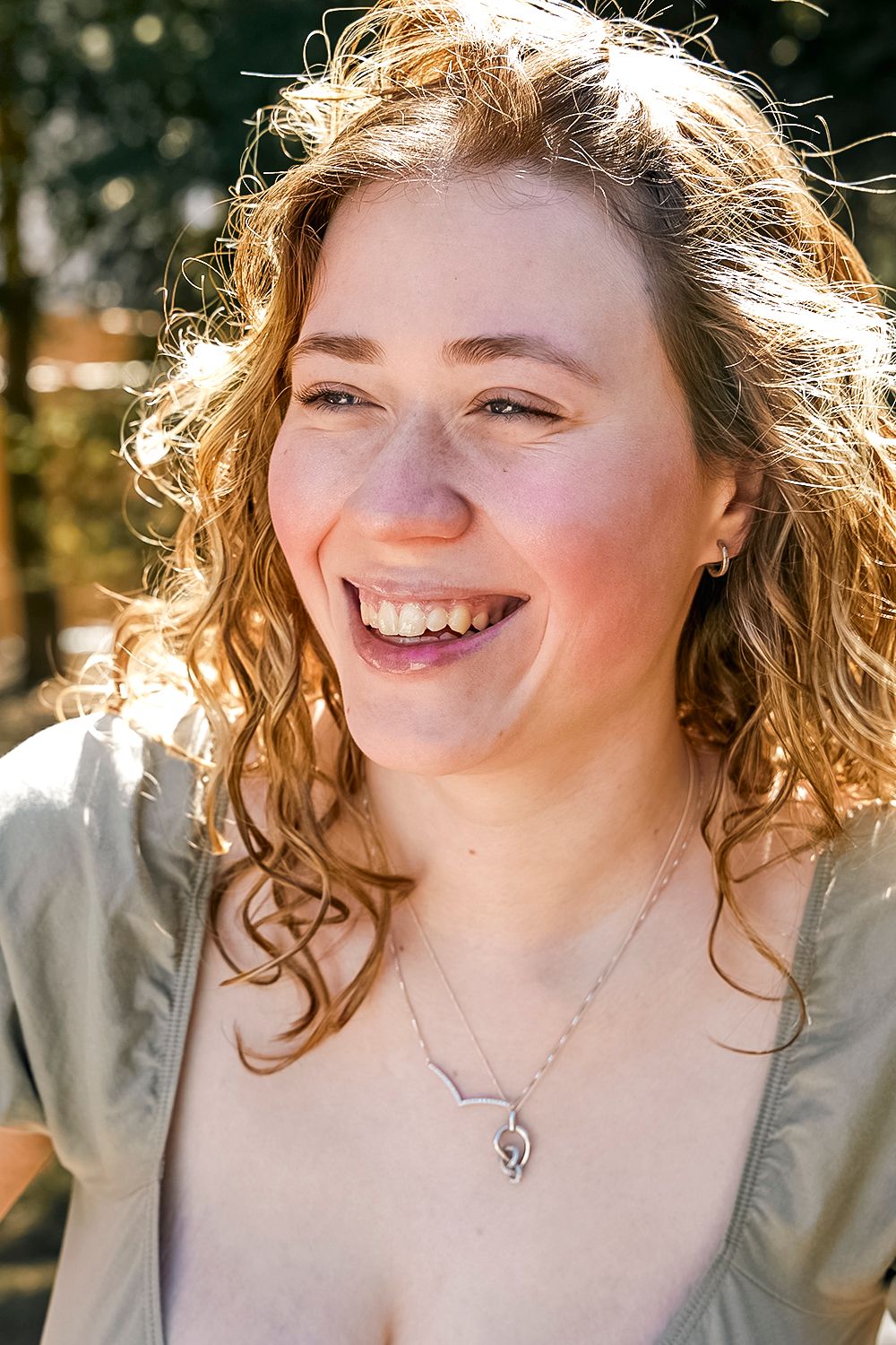 Natural sunlit portrait of someone with curly hair wearing delicate necklace outdoors.
