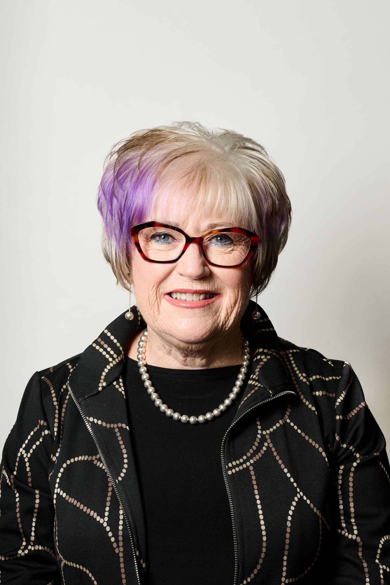Professional portrait showing someone wearing glasses and a black sweater with pearl necklace against white background.