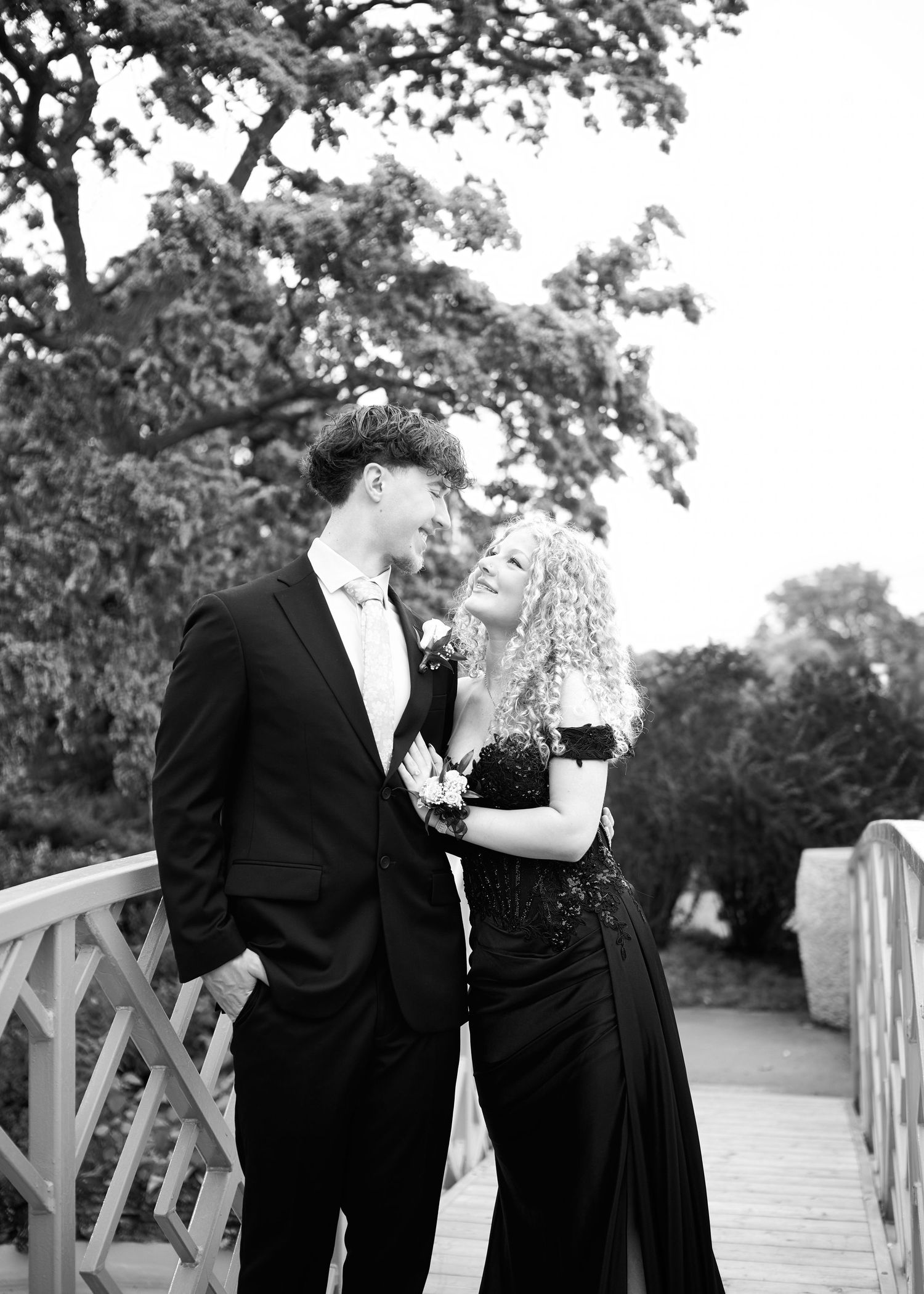 Black and white photo of a young couple in formal attire on a bridge, smiling at each other outdoors.