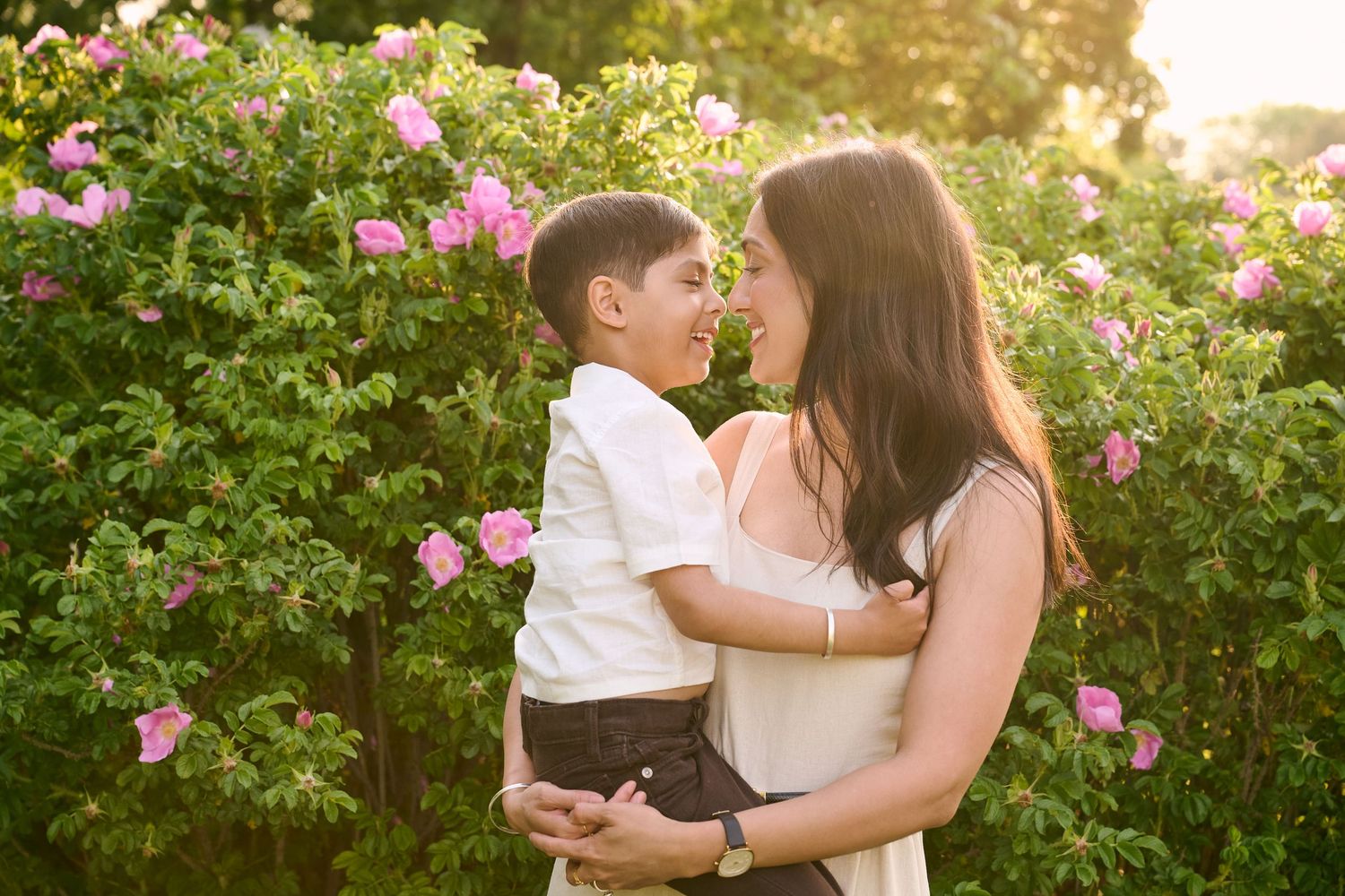A mother holds her young son among pink blooming roses in a sunlit garden, sharing a tender nose-to-nose moment.