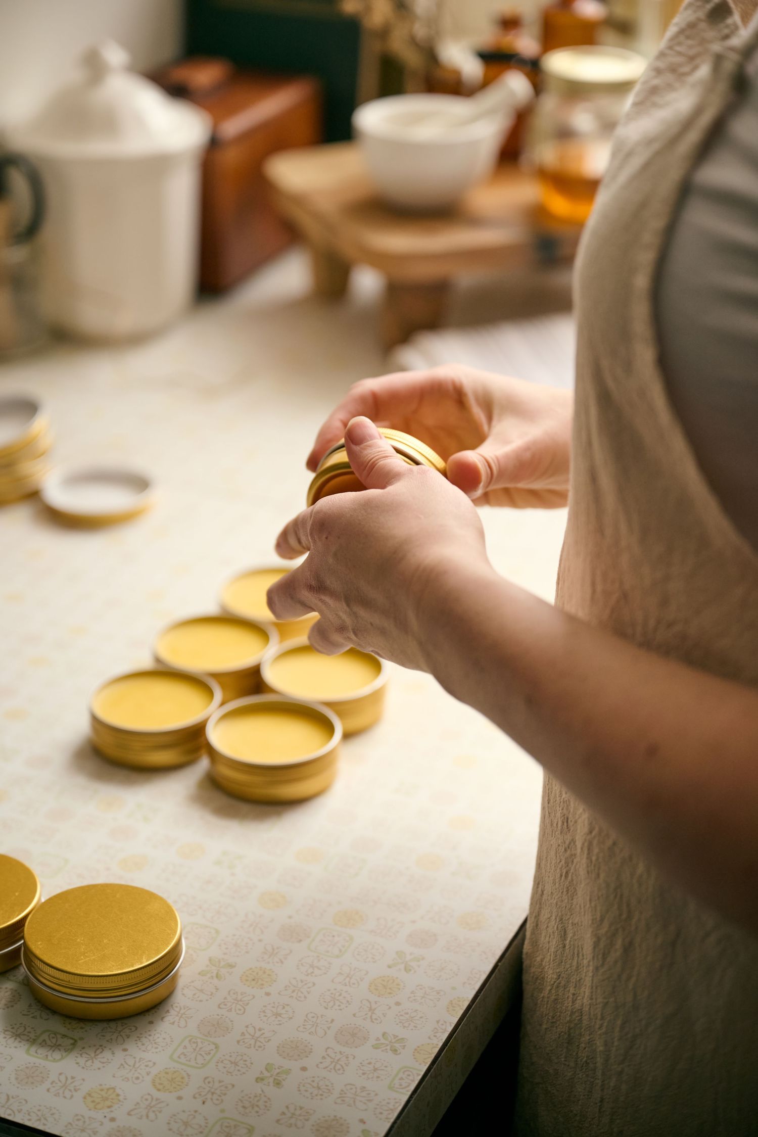 Hands preparing small yellow jar lids on a white countertop with teapots and kitchen items in the background.