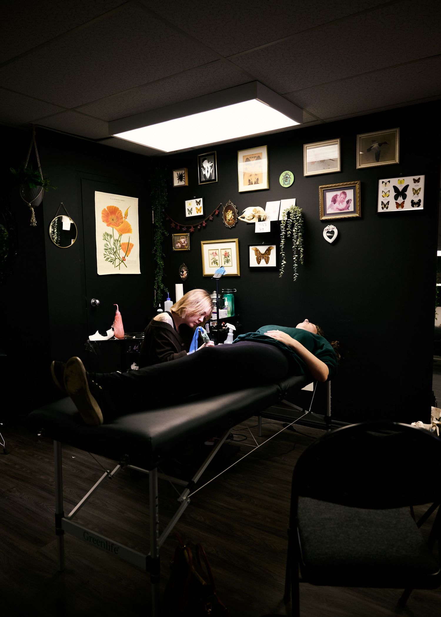 A tattoo artist working in a dark studio with framed artwork on black walls and bright overhead lighting.