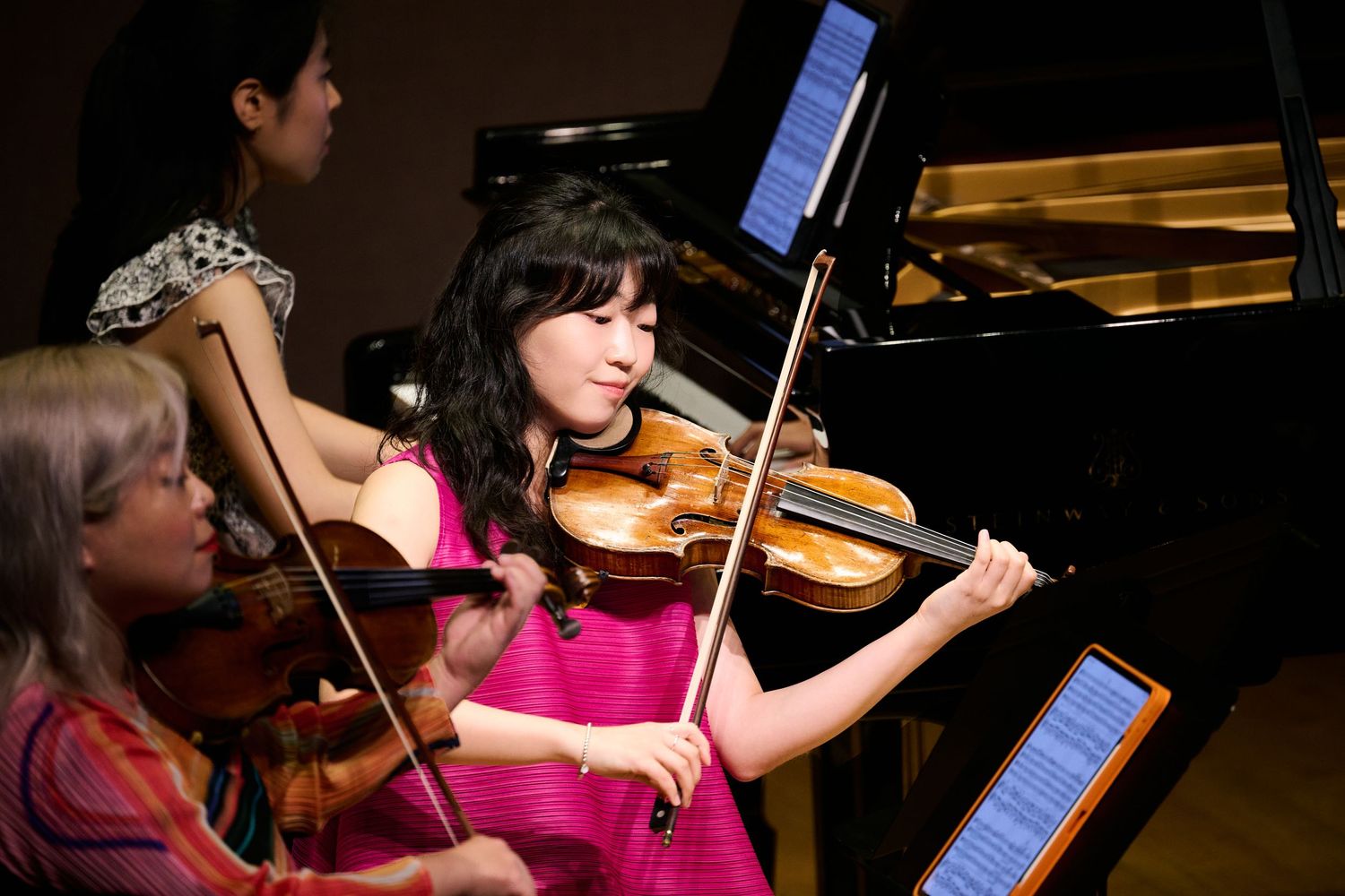 Two female violinists perform chamber music on stage near a grand piano during a classical music concert.