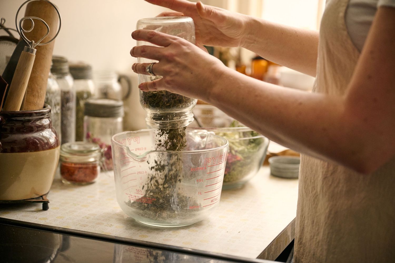 Hands sprinkle dried herbs into a glass jar on a kitchen counter with other cooking ingredients and supplies nearby.