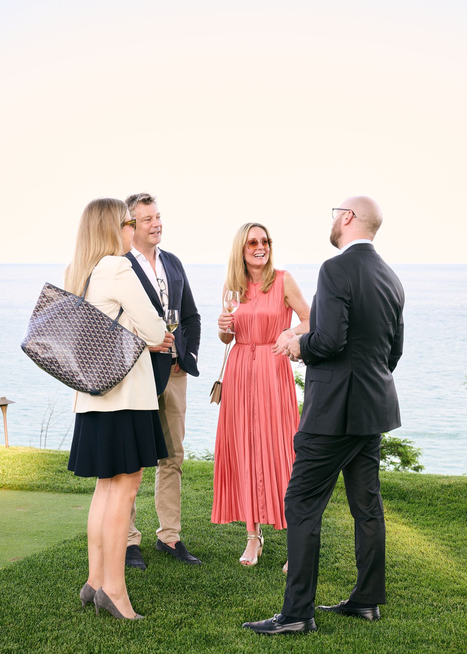 Four elegantly dressed guests socializing outdoors on a lawn with champagne glasses near an ocean view.
