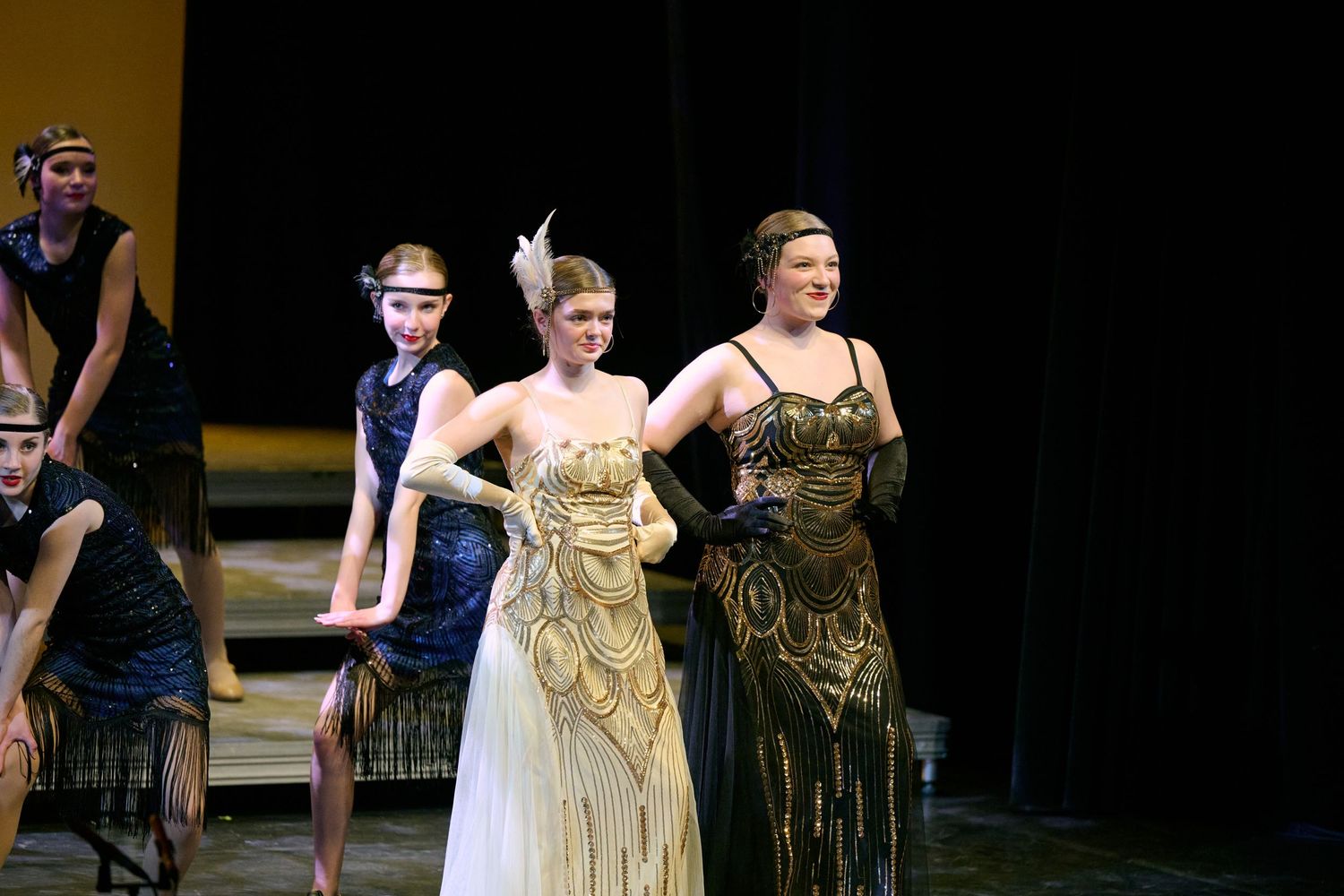 Performers in elegant gold and olive evening gowns stand on stage during a formal musical performance.