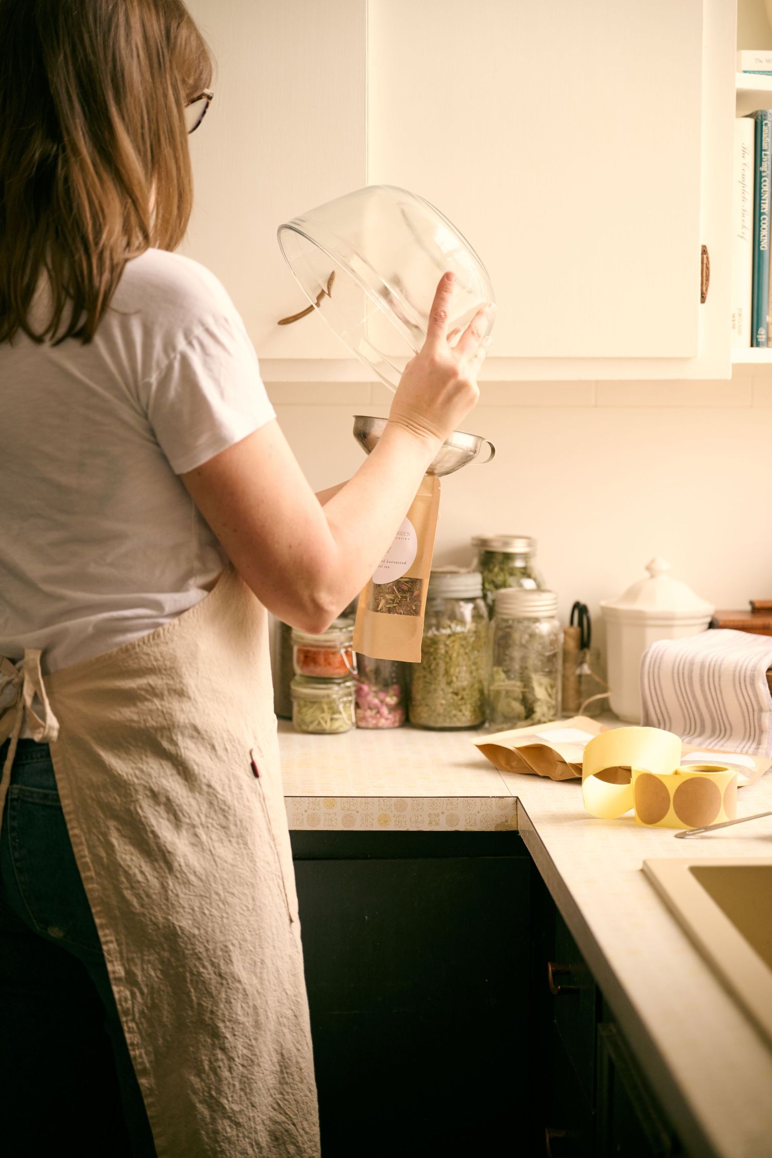 Person in white shirt and apron cooking in kitchen with glass jars of ingredients and measuring cups on counter.