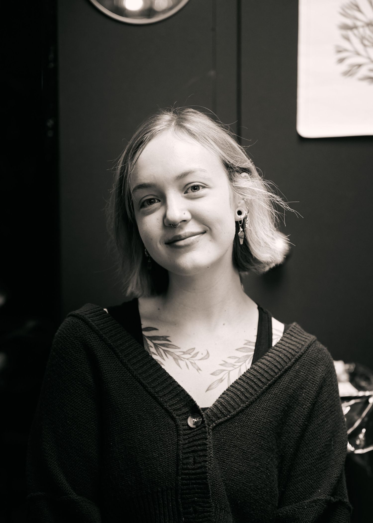 Black and white portrait photograph showing someone in a dark cardigan sweater with tattoos and a gentle smile.