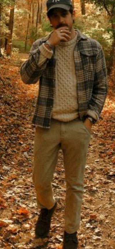 Person in plaid jacket and Yankees cap poses in autumn forest with fallen leaves on ground.
