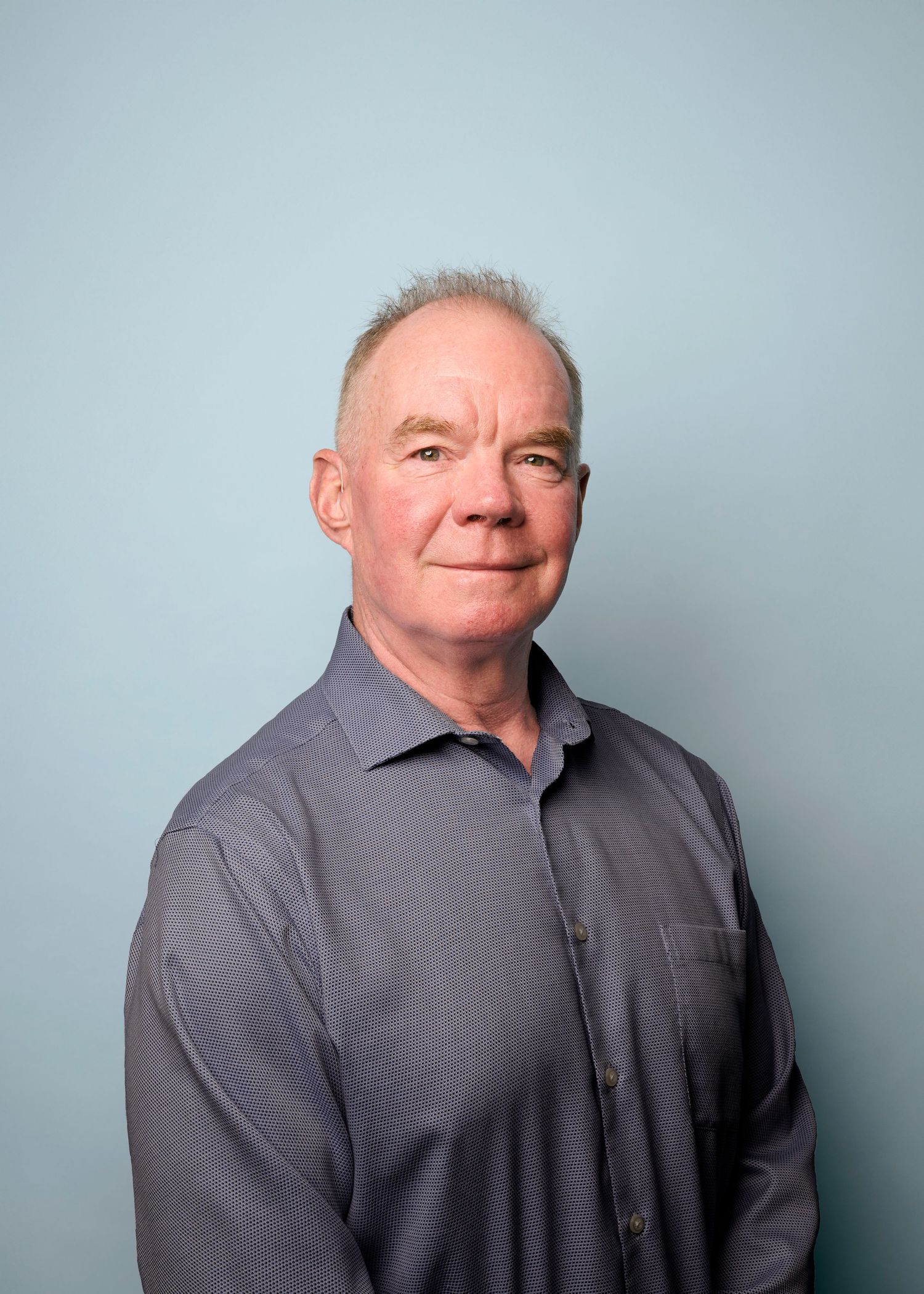 Professional headshot portrait of a person in a gray button-up shirt against a light blue background.