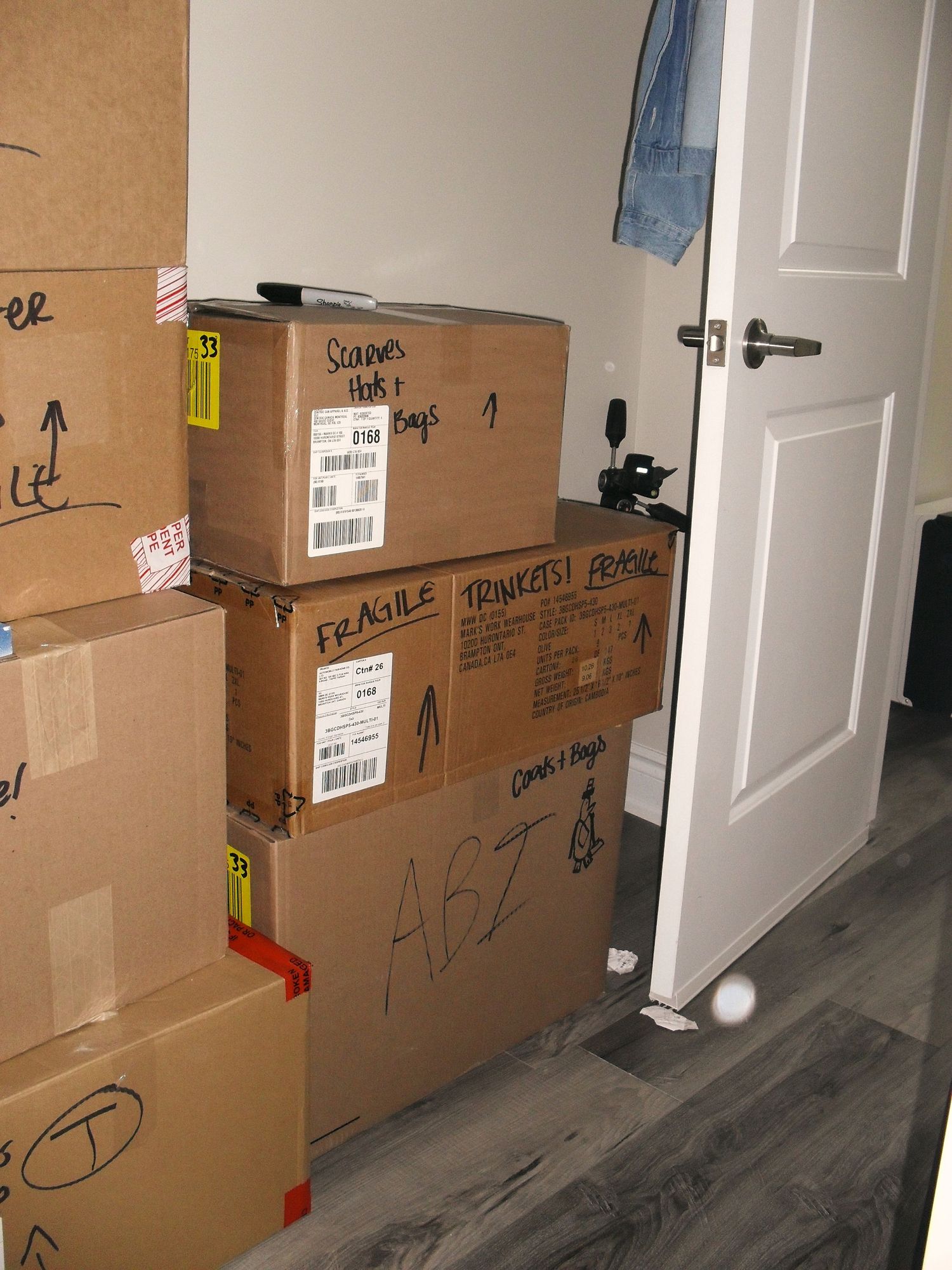 Several stacked cardboard moving boxes with shipping labels and handwritten markings against a white door and grey floor.