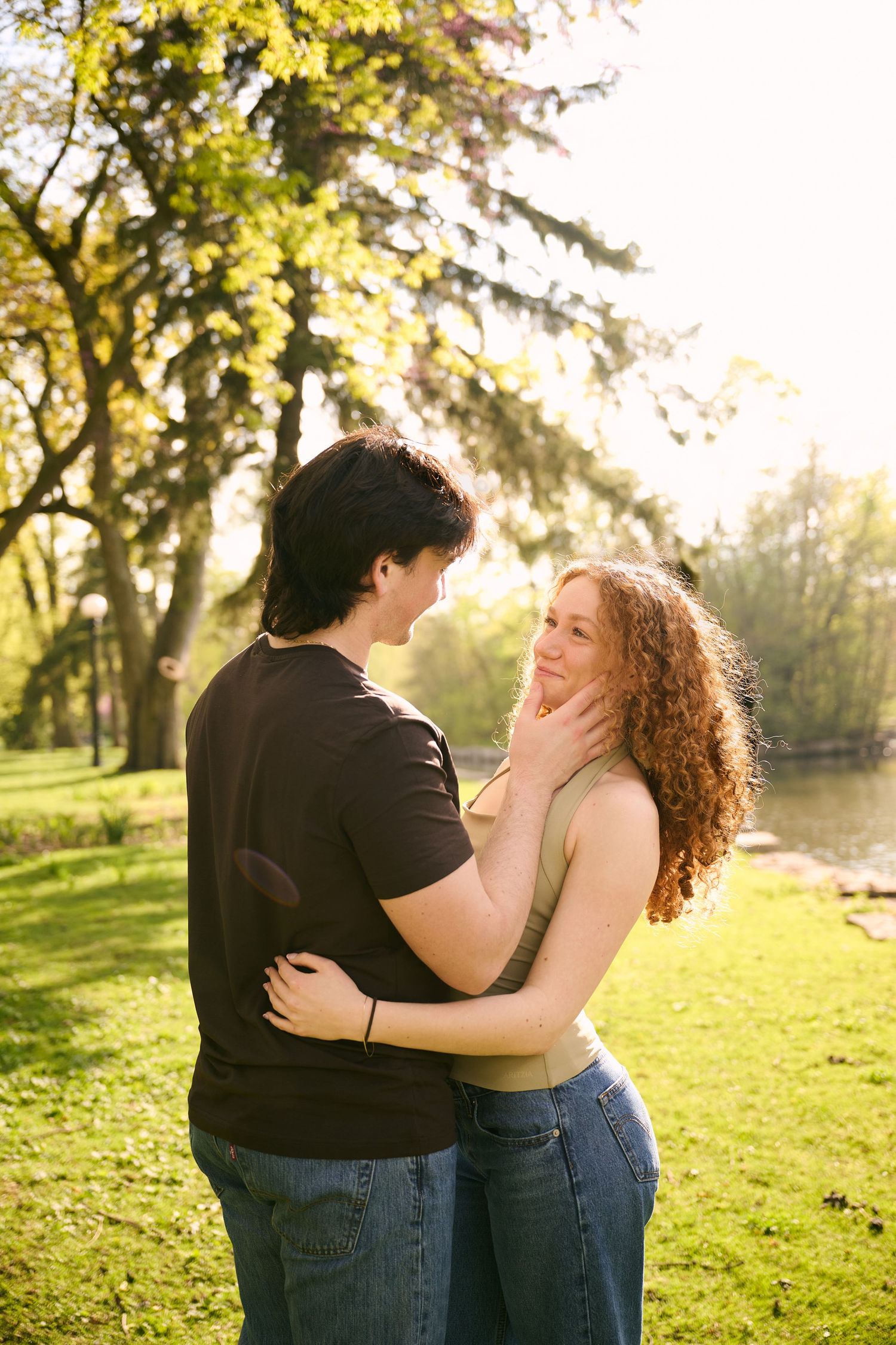 Romantic couple embracing in a sunny park, woman with curly red hair smiling as man holds her face tenderly.
