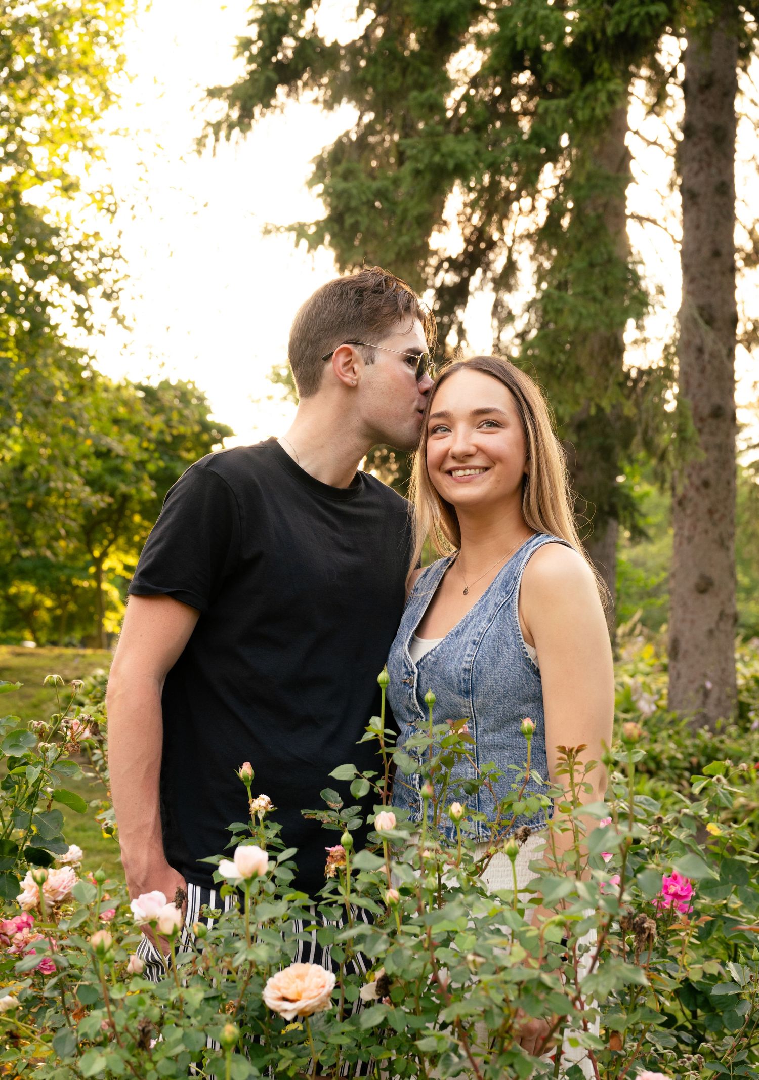 A romantic couple shares a tender moment among blooming roses in a sunlit garden setting.