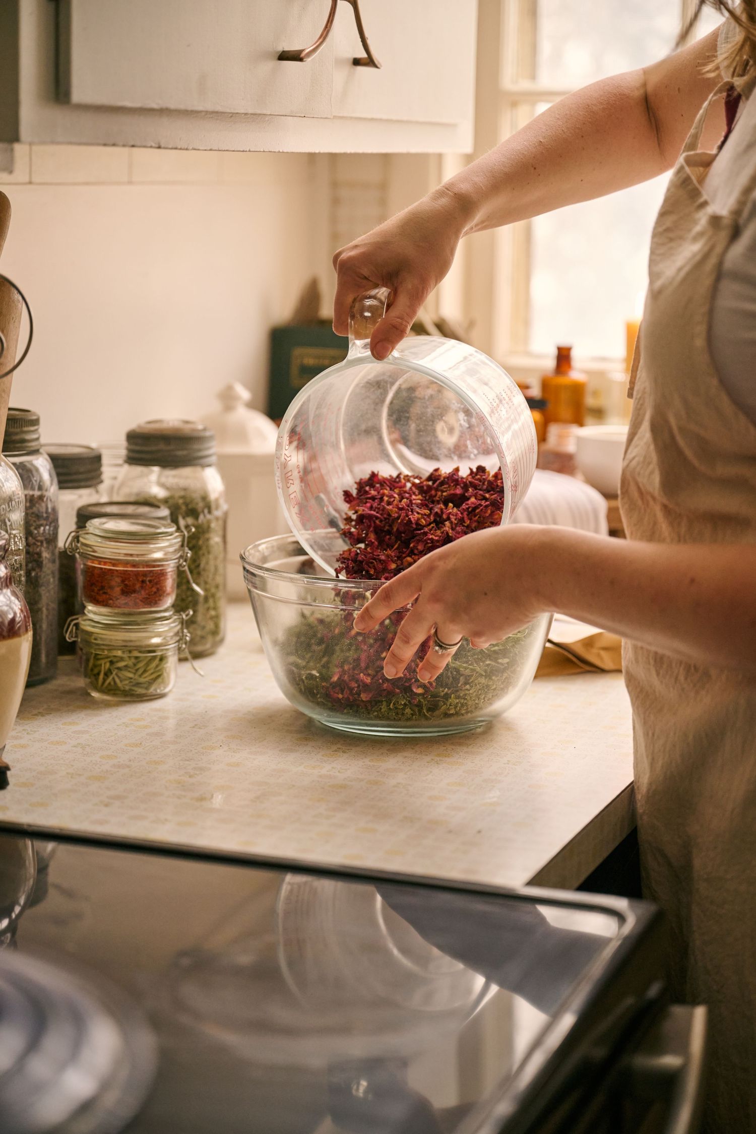 Hands adding dried herbs to a glass mixing bowl in a sunlit kitchen.