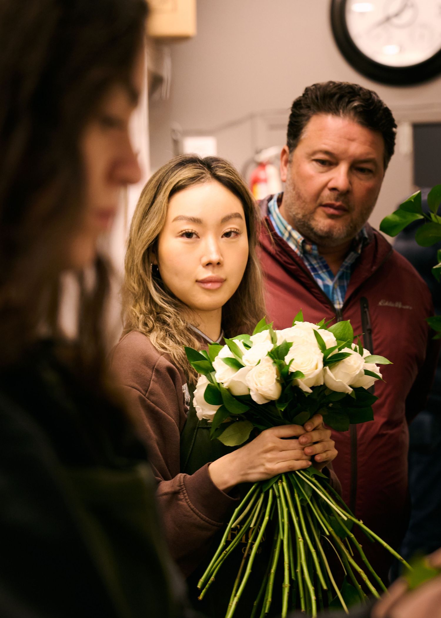 A group of people in a dimly lit room examines a bouquet of white flowers held by someone in a brown sweater.